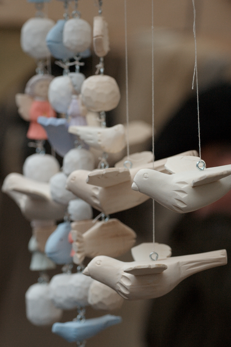 Close-up of multiple beige and white clay bird ornaments from Kazjukas 2012 hanging on strings. The birds are aligned in a row, appearing to be in mid-flight. The background is blurred, showing hints of other objects in light blue, pink, and gray, but details are indistinct.