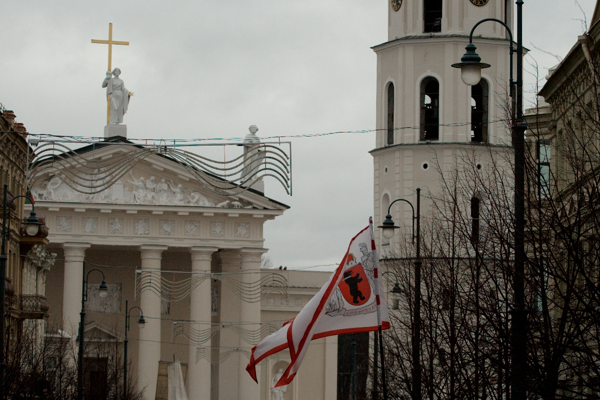 A white church with a tall bell tower featuring a clock and arched windows stands under a cloudy sky, reminiscent of Kazjukas 2012. The church has a pediment with statues and a cross on top. Two red and white flags with an emblem wave in the breeze, while leafless trees and street lamps are visible in the foreground.