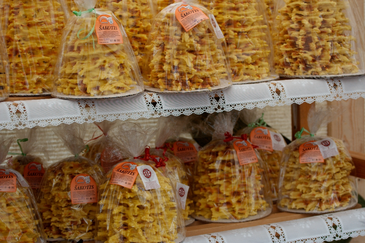 This image shows multiple traditional Lithuanian tree cakes, known as Šakotis, on display at Kazjukas 2012. The cakes are intricate and spiky, resembling small trees with yellowish-brown hues. They are packaged in clear plastic and have labels attached, placed on shelves lined with white lace cloth.