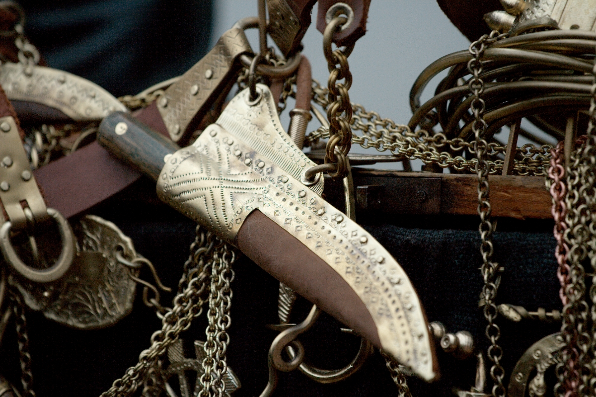 A decorative knife with an intricately engraved blade and handle, adorned with patterns and studded details, hangs among an assortment of chains and other metallic objects. The knife, reminiscent of a 2012 Kazjukas design, has a partially visible wooden handle and seems part of an elaborate collection.