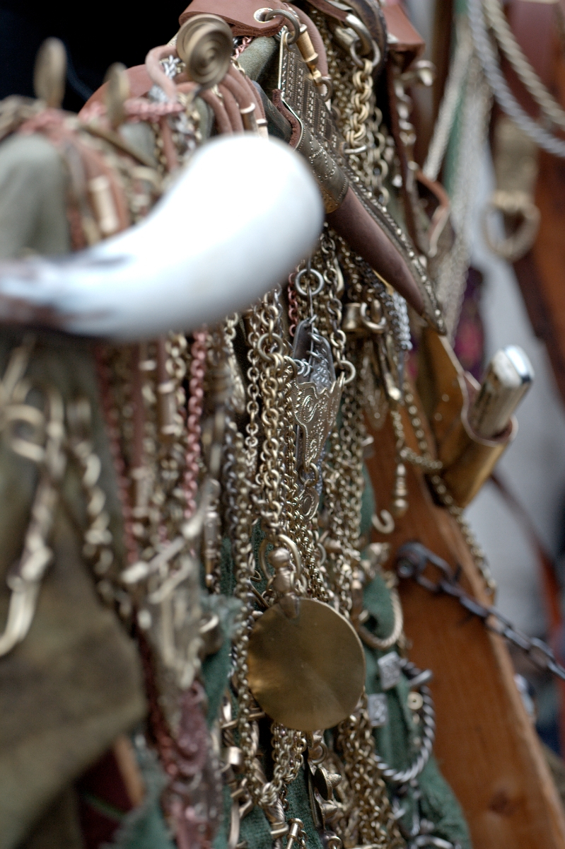 A close-up image of an assortment of brass chains, ornate keys, and various metal trinkets hanging on a green fabric. The items include a white horn and a leather sheath containing a small dagger. The scene appears cluttered, showcasing intricate, vintage items reminiscent of Kazjukas 2012.