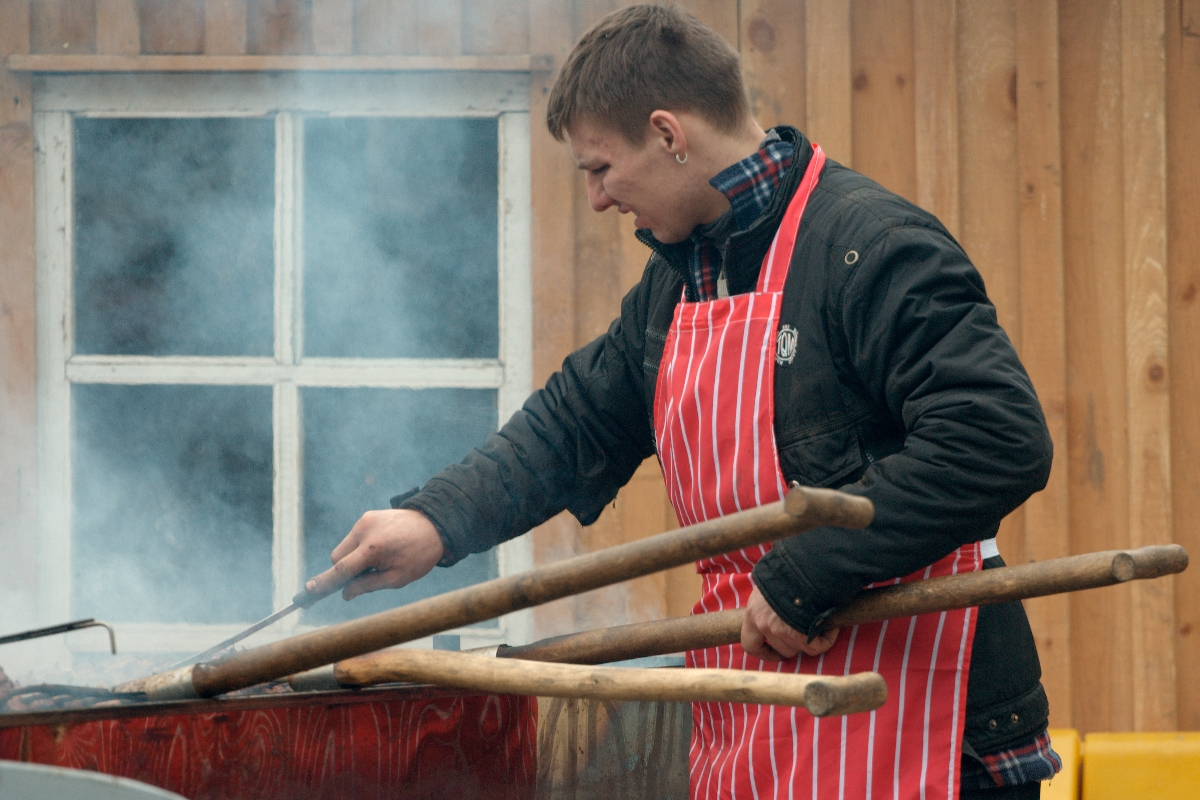 A person wearing a red and white striped apron and a black jacket is grilling food on a barbecue, reminiscent of the scene from Kazjukas 2012. They are holding large wooden tongs and turning food on the grill with a metal spatula. Steam or smoke rises from the barbecue, with a wooden building and window in the background.