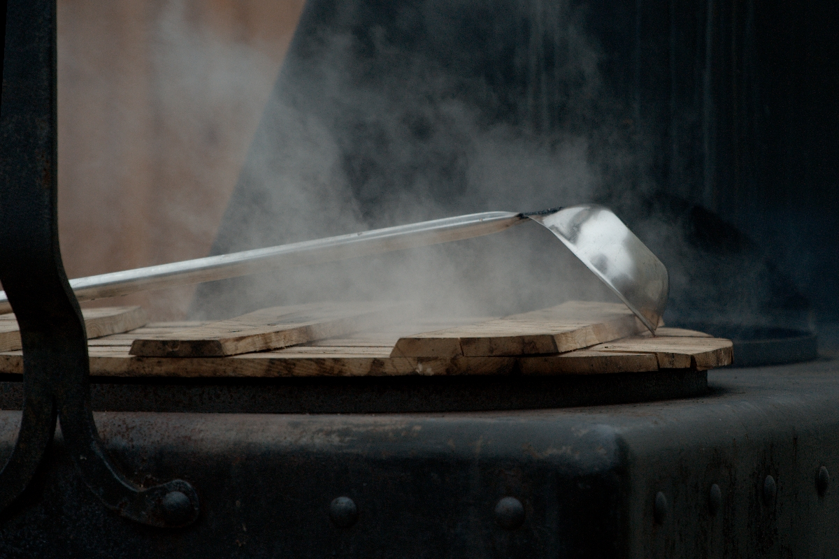 Close-up of a steel ladle resting on a wooden surface, with steam rising up, suggesting it's hot. The dimly lit background emphasizes the steam and the contrast between the metal ladle and wood below. The scene, evoking a 2012 Kazjukas aesthetic, conveys a rustic or industrial cooking environment.