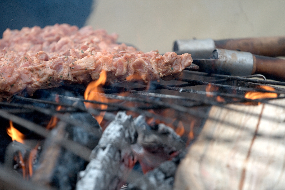 A close-up of skewered pieces of marinated meat cooking over a charcoal grill with visible flames and embers, reminiscent of Kazjukas 2012. The chicken appears well-seasoned, turning golden from the heat. The darkened grill rods hint at frequent use, adding to the warm, smoky ambiance.