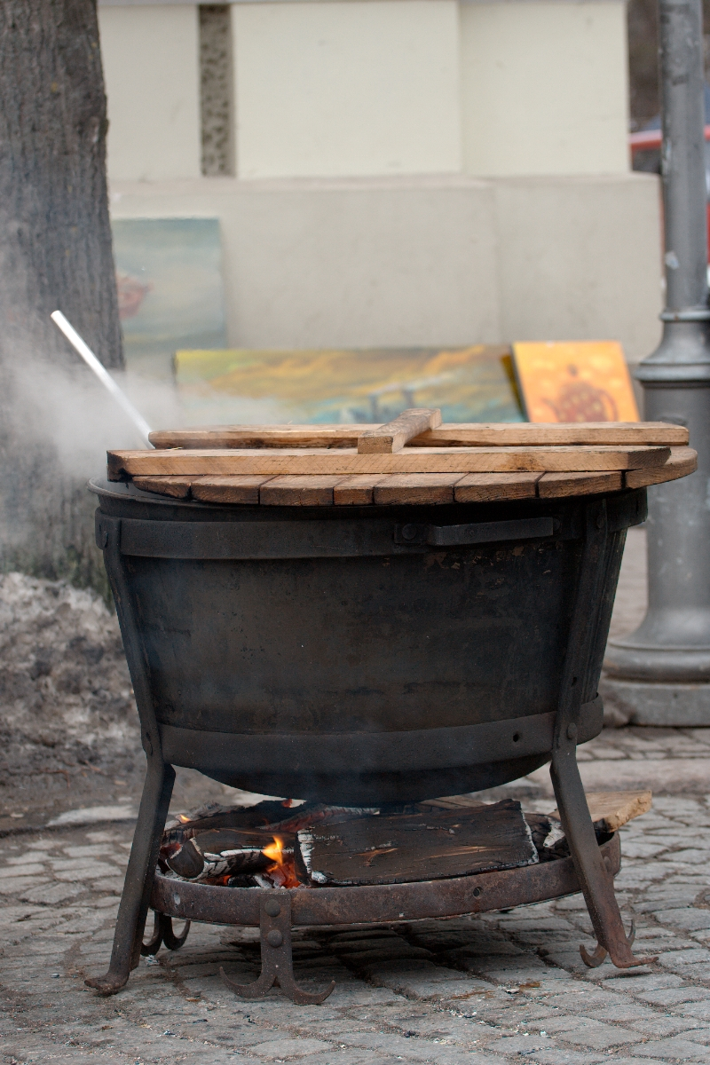 A large black pot labeled "Kazjukas 2012" sits on an outdoor wood-fired stove with a wooden lid partially covering it. The stove has a metal frame and visible flames below, indicating it is actively in use. In the blurred background, there are some paintings and a tree trunk. A metal lamp post is also visible.