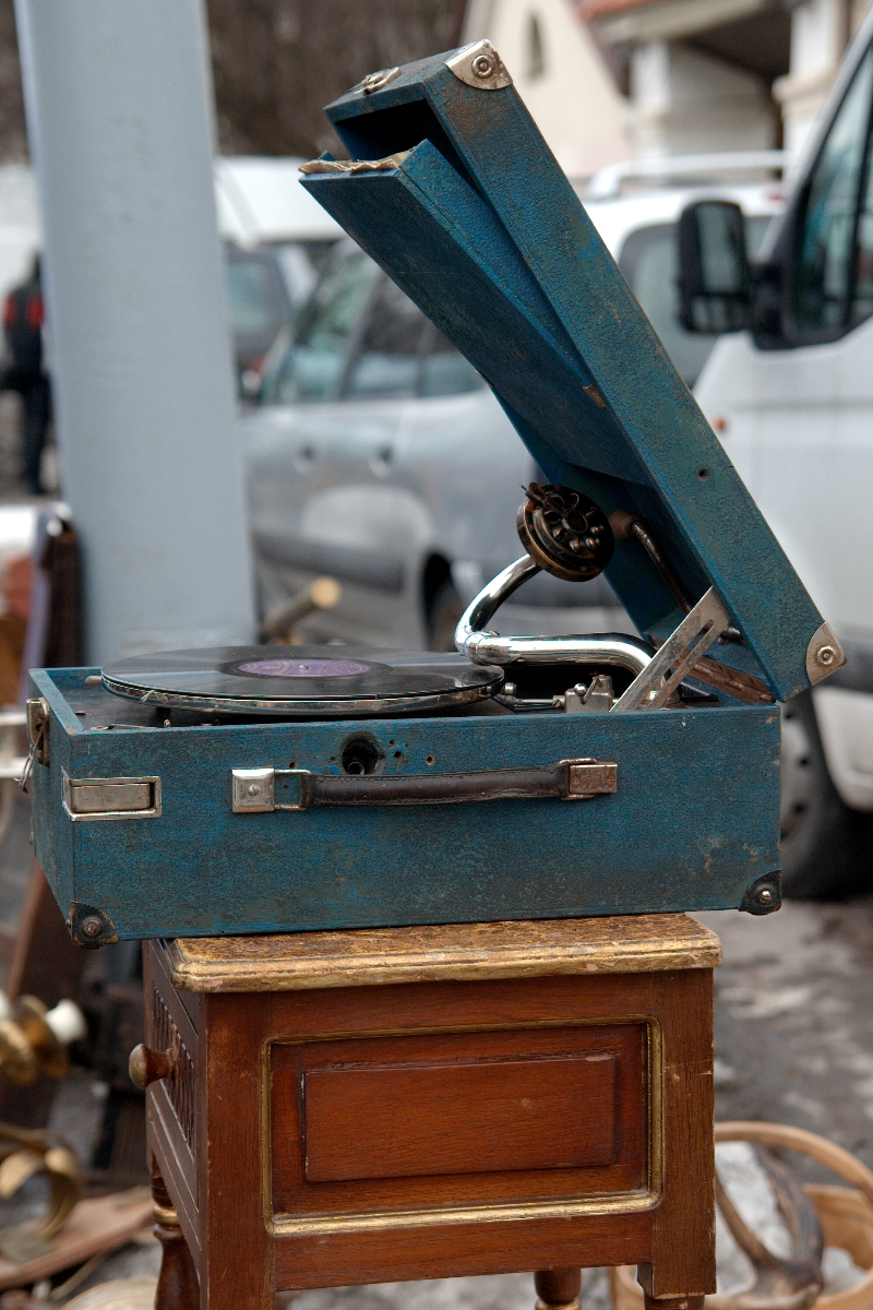 A vintage blue gramophone with a record on the turntable sits on a small brown wooden table. The gramophone's lid is open, revealing the horn and tonearm. In the background, a white van and another vehicle suggest an outdoor setting like Kazjukas 2012 flea market or street sale.