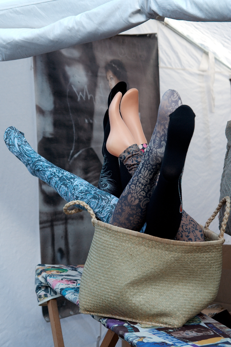 A woven basket tipped over reveals several mannequin legs protruding in different directions. The legs are adorned with various tights and stockings, featuring patterns such as floral, paisley, and solid black. The scene is set against a backdrop with a partially visible image and white fabric, reminiscent of Kazjukas 2012.
