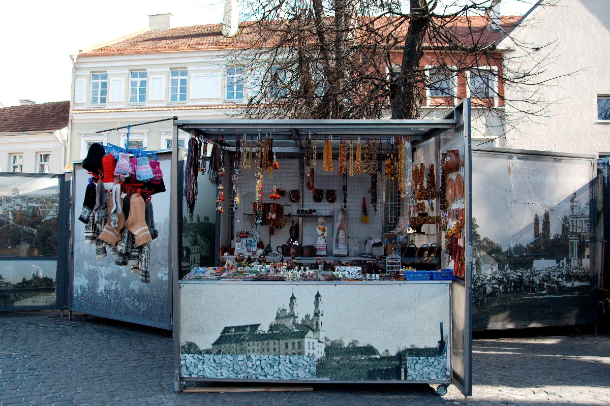 A street-side kiosk displays various amber jewelry, leather crafts, traditional hats, and Kazjukas souvenirs. It has a backdrop depicting a historic cityscape from 2012. There is a stone-paved street in front, and the kiosk is set against a background of buildings and bare trees.