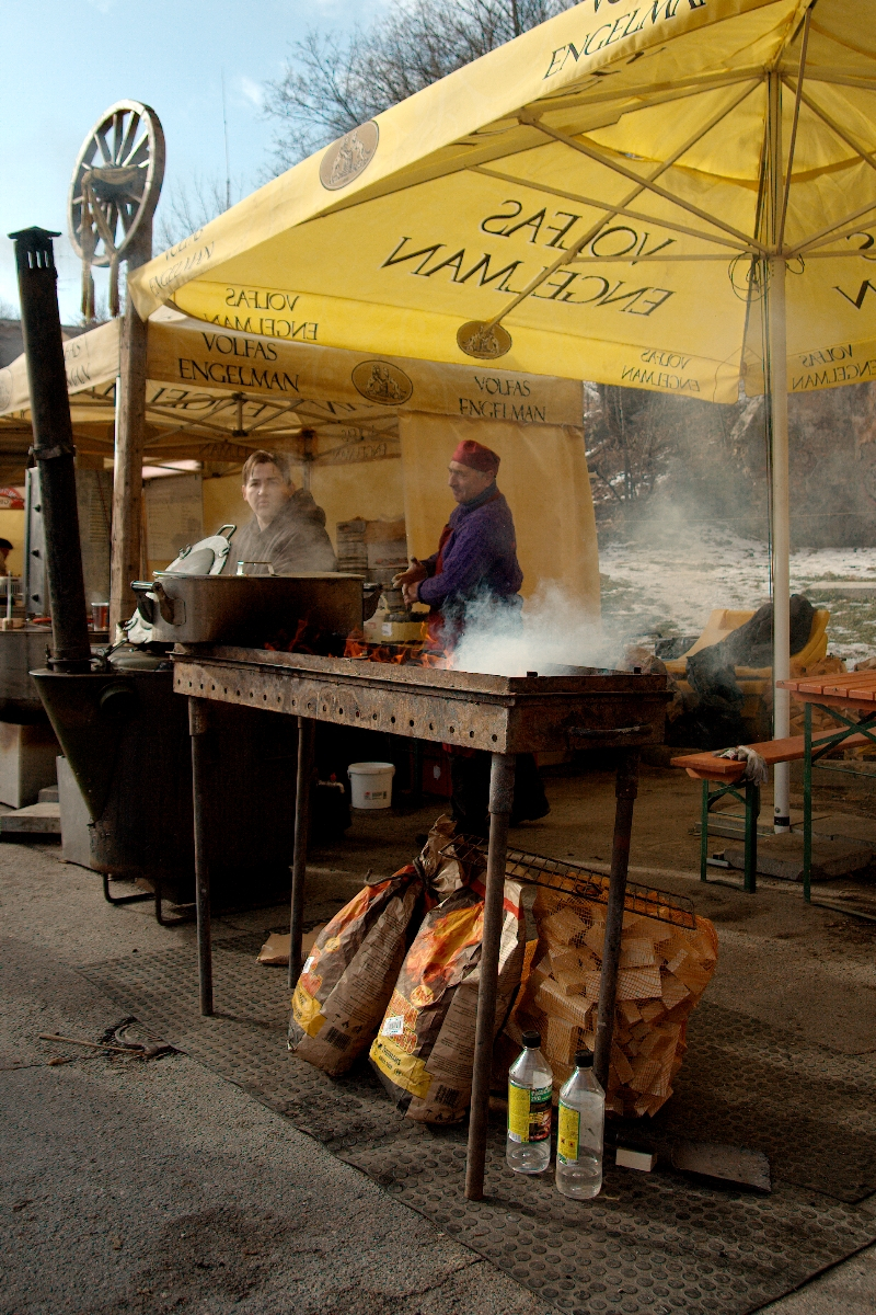 Two people are cooking under large yellow Volfas Engelman-branded umbrellas. One person is at a grill with smoke rising and the other stands behind a table. Bags of firewood and two water bottles are in front of the grill. The outdoor setting has the cooks in winter clothes, reminiscent of Kazjukas 2012.