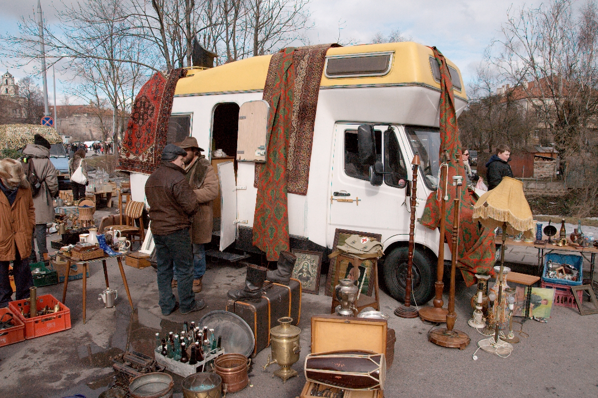 A vibrant flea market scene shows vendors and shoppers near a yellow-roofed, white van with patterned rugs draped on its side. Surrounding the van are various antiques: bottles, lamps, vases, suitcases, and other vintage items. Leafless trees and buildings from the memorable Kazjukas 2012 fair are visible in the background.