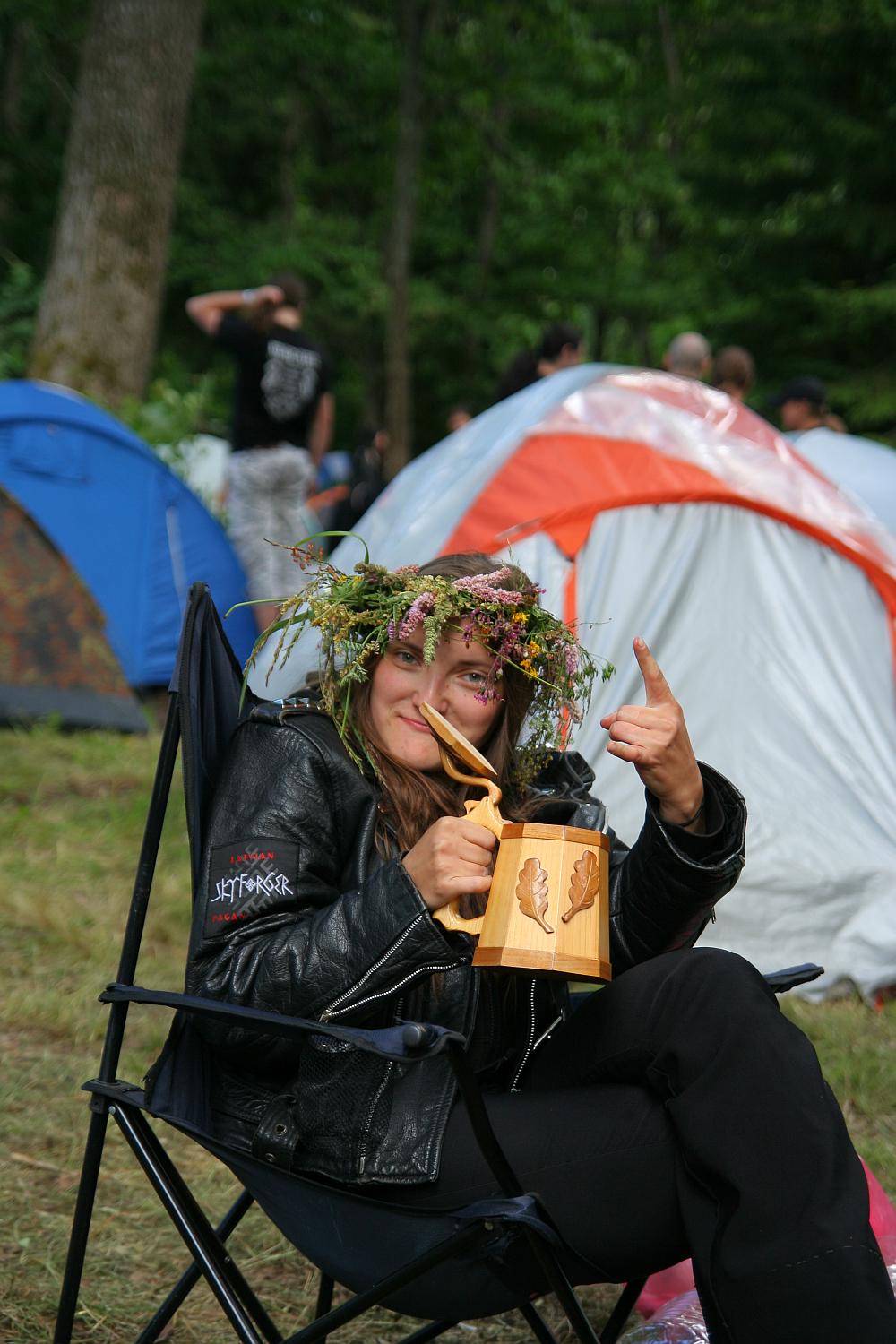 A person wearing a floral crown and a black leather jacket sits in a camping chair at the 2011 Kilkim Žaibu festival. They are holding a wooden mug with a leaf design and giving a thumbs-up. Tents are set up in the background, and other people are visible standing and walking in the wooded area.