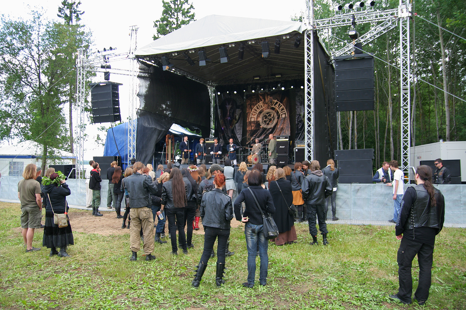 A small crowd of people, many dressed in black leather jackets, stand and watch a rock band performing on an outdoor stage at the 2011 Kilkim Žaibu festival. The stage has large speakers and lighting rigs. Trees and grass surround the area, creating a natural setting. Some people are taking photos.
