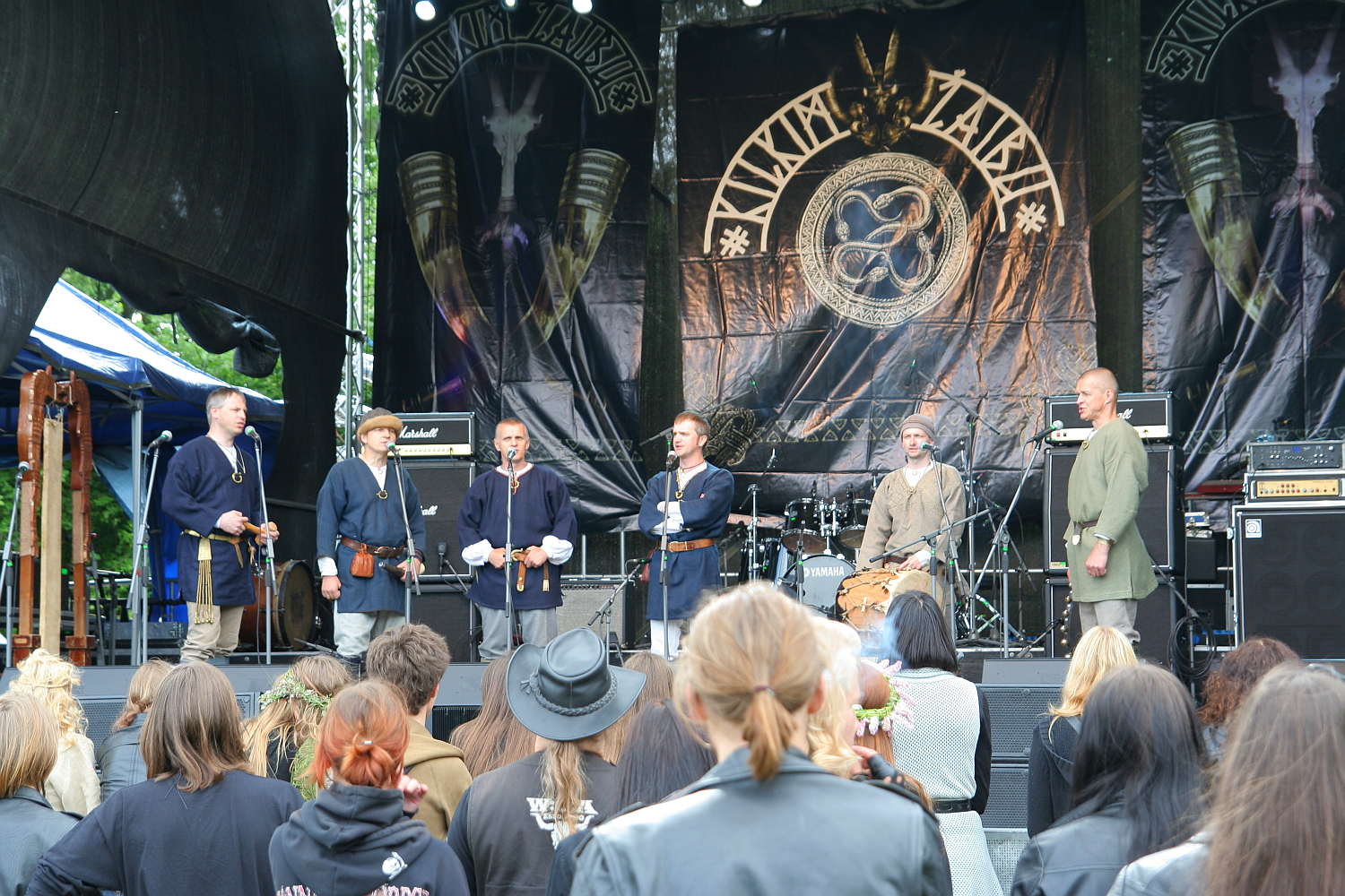 A group of six men dressed in medieval-style clothing stands on a stage surrounded by instruments and audio equipment. They face an audience dressed in casual, modern attire. The backdrop features a large logo and the text "Antiytila" with Viking symbols, echoing the spirit of Kilkim Žaibu 2011. Surrounding trees are partially visible.