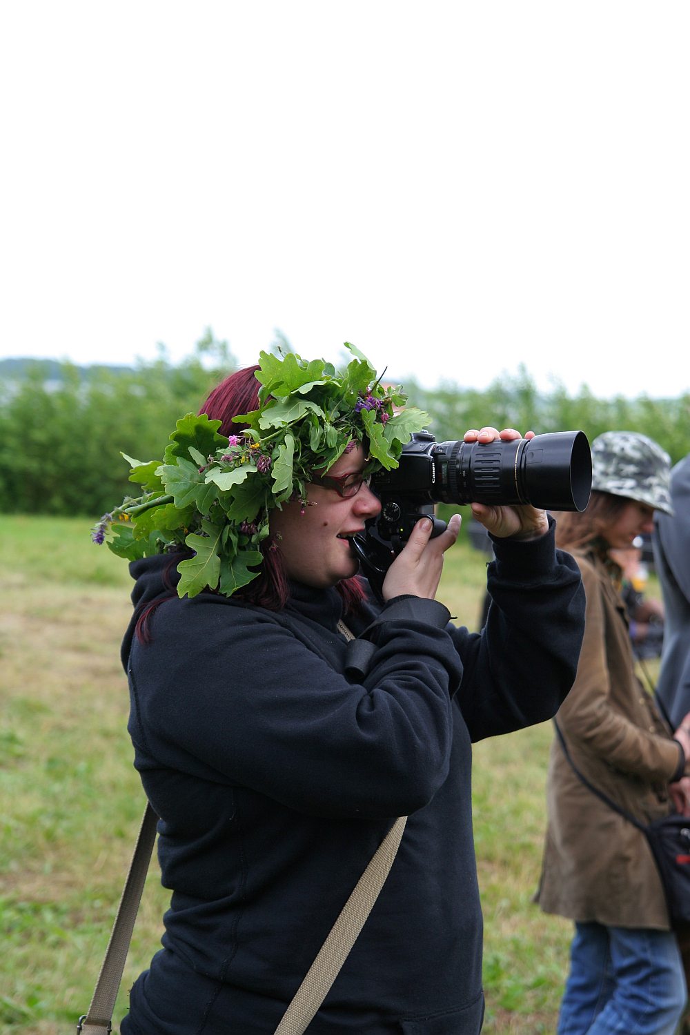 A person with dark hair, wearing a black hoodie and an elaborate green leafy crown, looks through a large camera lens in an outdoor field during the 2011 Kilkim Žaibu festival. Other individuals are blurred in the background, suggesting a group event in a natural setting. The sky appears overcast.