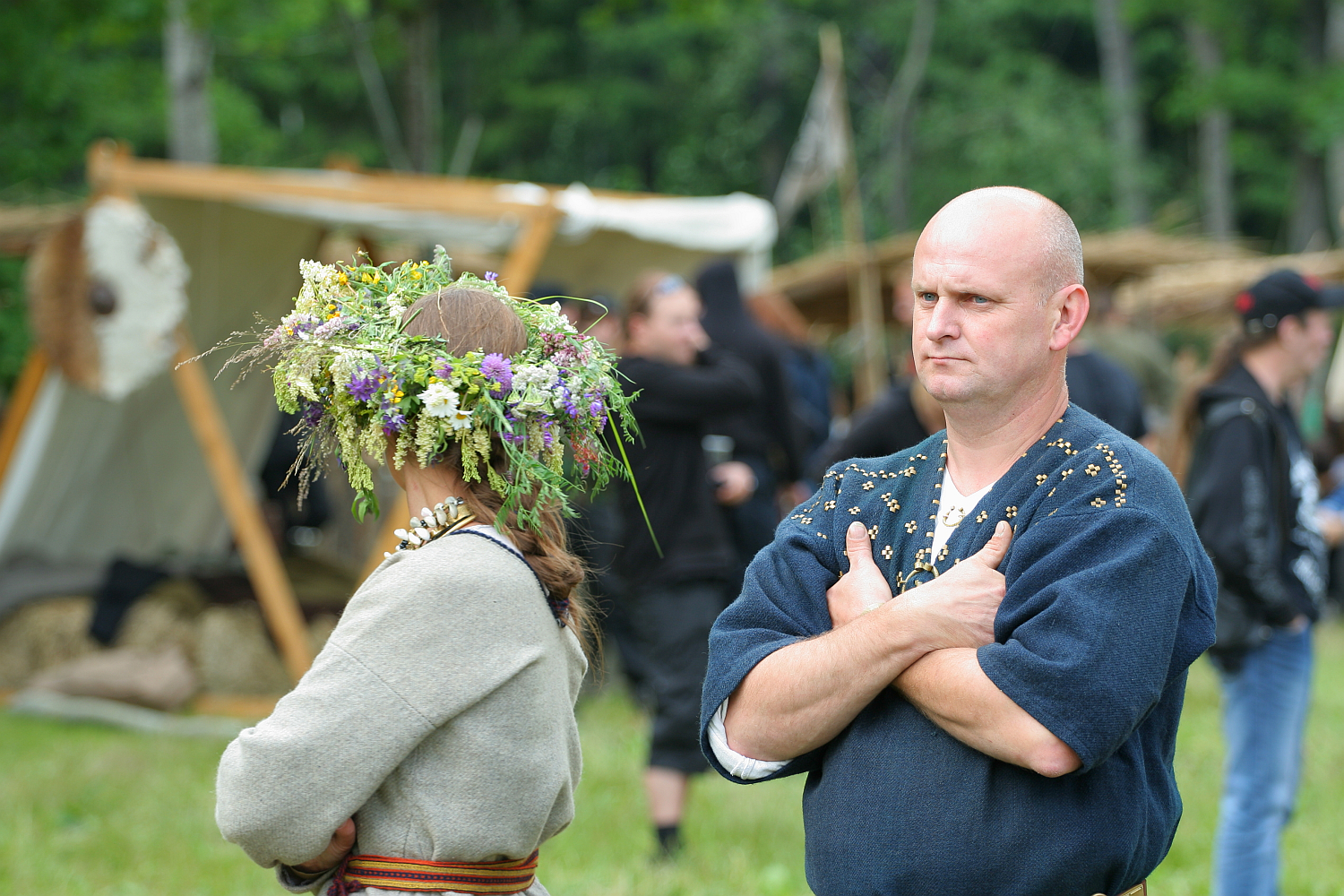 Two individuals participate in an outdoor historical reenactment at Kilkim Žaibu 2011. The woman on the left wears a flower crown and a traditional dress, her back to the camera. The man on the right stands with arms crossed, wearing traditional attire. A rustic tent and other participants are visible in the background.