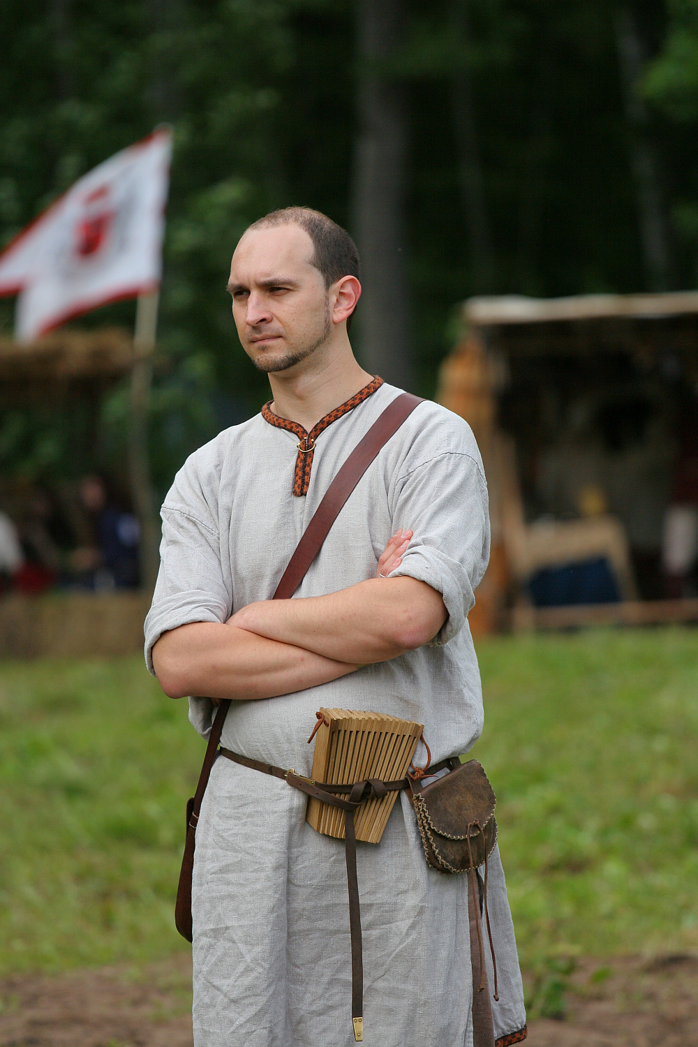 A man dressed in medieval attire stands with folded arms. He wears a light gray linen tunic, a brown leather belt with a pouch, and a shoulder strap holding a wooden object. Behind him is a blurred background of greenery and a flag with a red emblem, reminiscent of Kilkim Žaibu 2011.