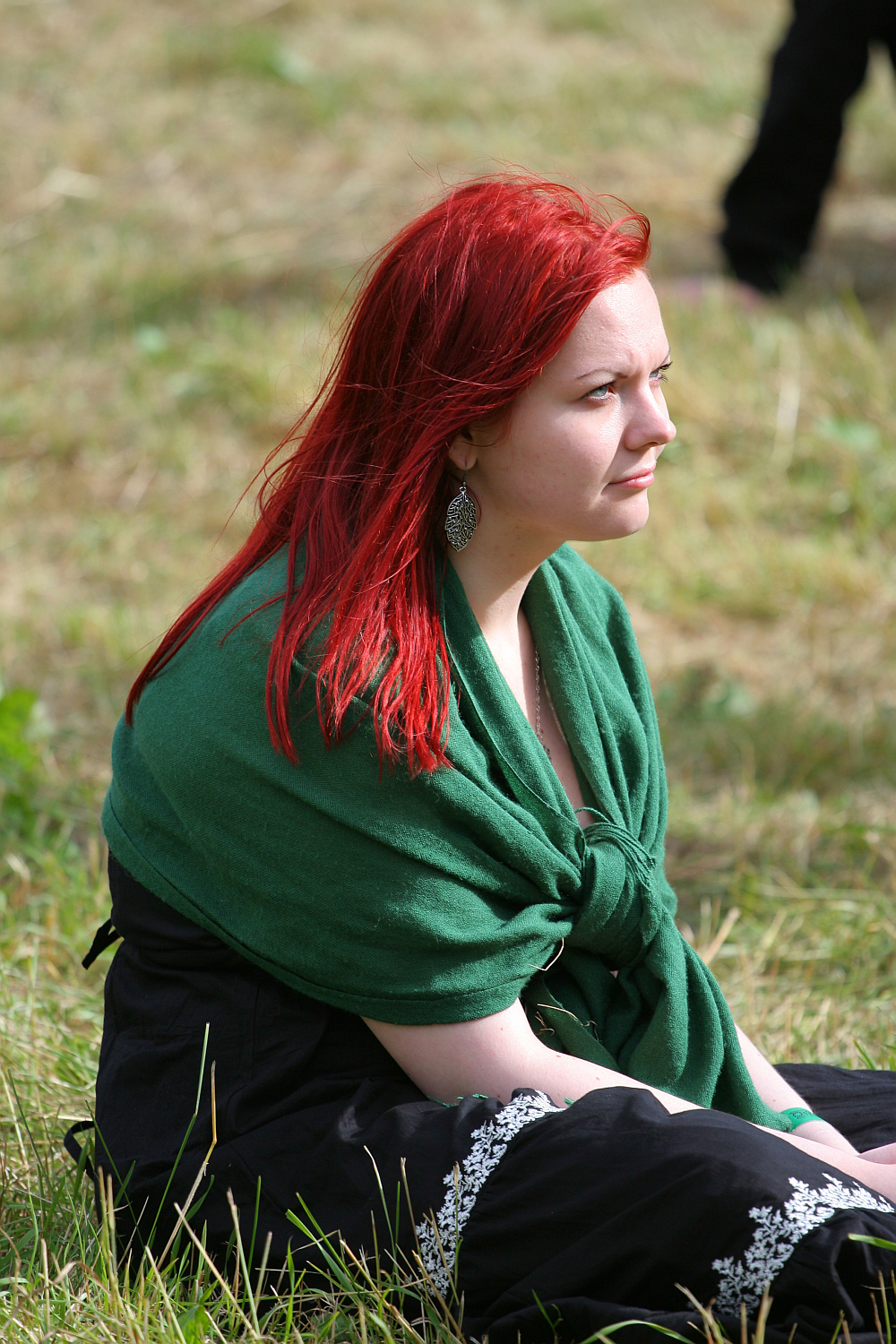 A red-haired woman wearing a green shawl over a black dress sits on grass at Kilkim Žaibu 2011. She looks towards the distance with a thoughtful expression. The background includes more grass and the legs of another person, which are partially visible, suggesting an outdoor environment, possibly a park.
