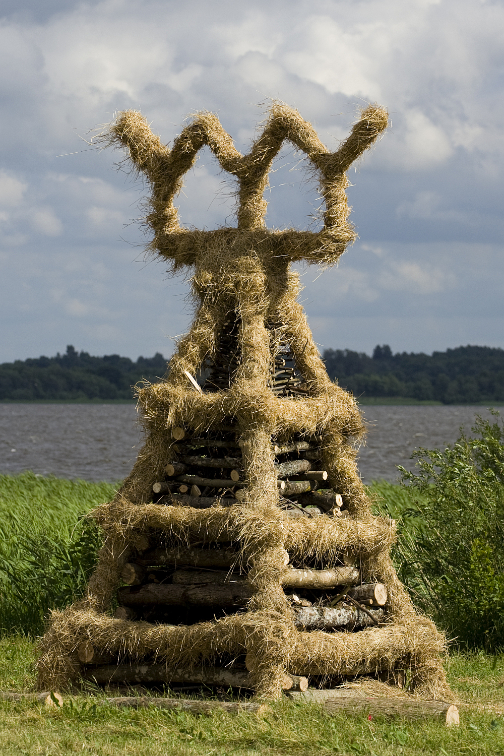 A large, intricately designed bonfire structure from the 2011 Kilkim Žaibu festival stands proudly on a grassy area near a body of water. Made of stacked logs and covered in straw, the conical structure features three prominent straw arches at the top, resembling a crown. Trees and a cloudy sky provide a picturesque backdrop.