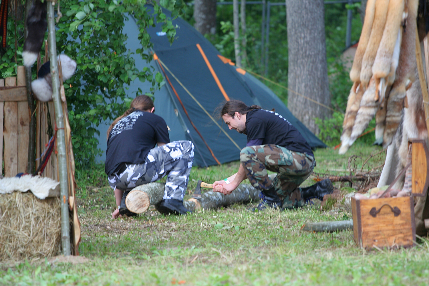 Two people in camo pants and black shirts are kneeling on the ground, chopping a log with an axe in a forested campsite during Kilkim Žaibu 2011. A tent is set up in the background, alongside a pile of fur pelts and wooden structures. The scene is set outdoors with green grass and trees visible.