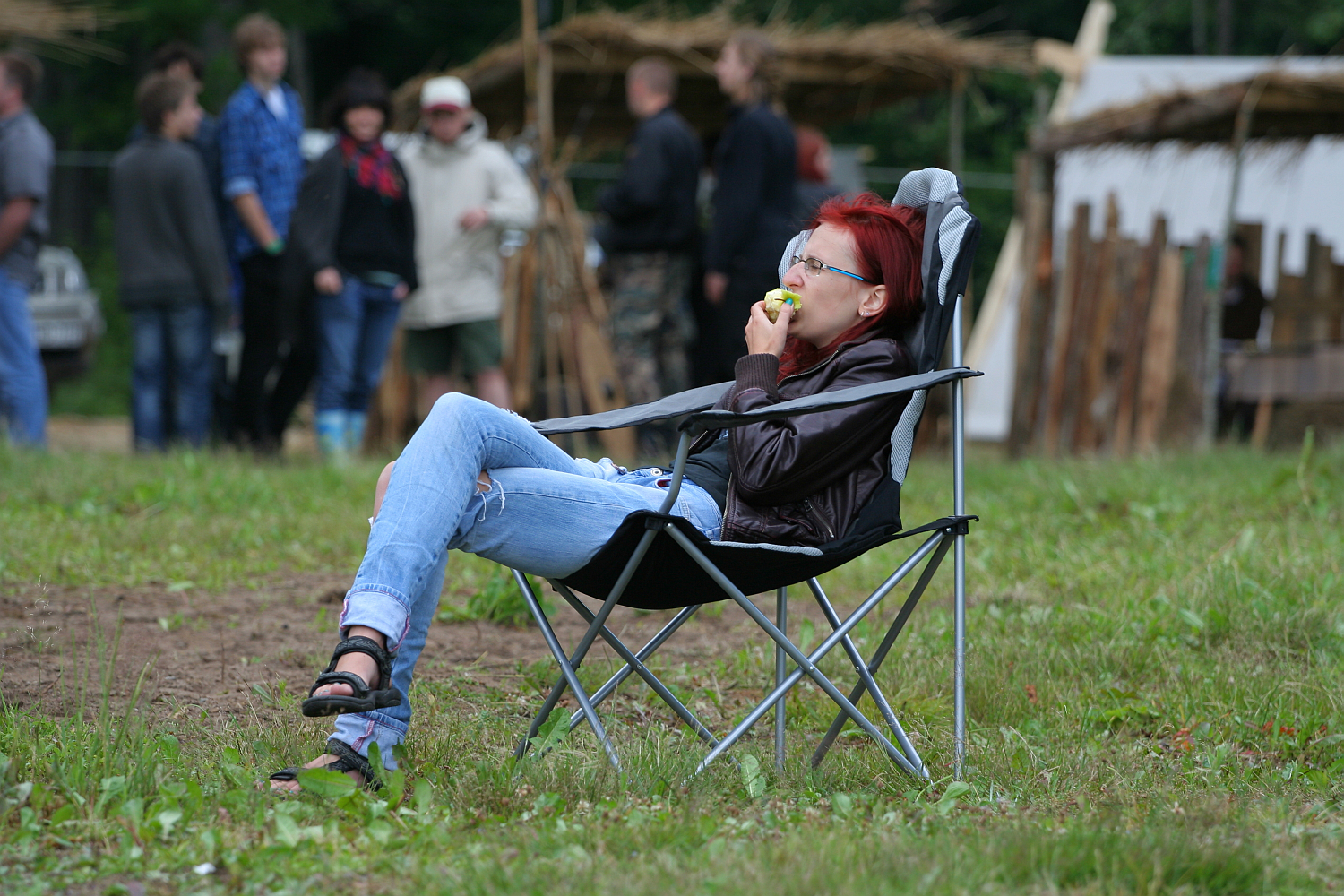 A person with short red hair and glasses relaxes in a black folding chair on a grassy field during Kilkim Žaibu 2011, eating an apple. Wearing a leather jacket, blue ripped jeans, and sandals, they lean back comfortably. In the background, several people are standing and talking near rustic, thatched structures.