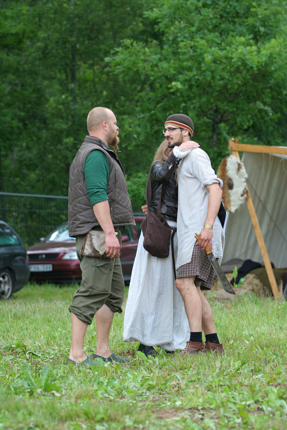 Three people in medieval attire converse outdoors at the 2011 Kilkim Žaibu festival. A bearded man in green and brown stands barefoot. A woman in a white dress and leather vest faces him, arm around a man in a headband, white tunic, and brown pants holding a sheathed sword. A tent and trees are in the background.