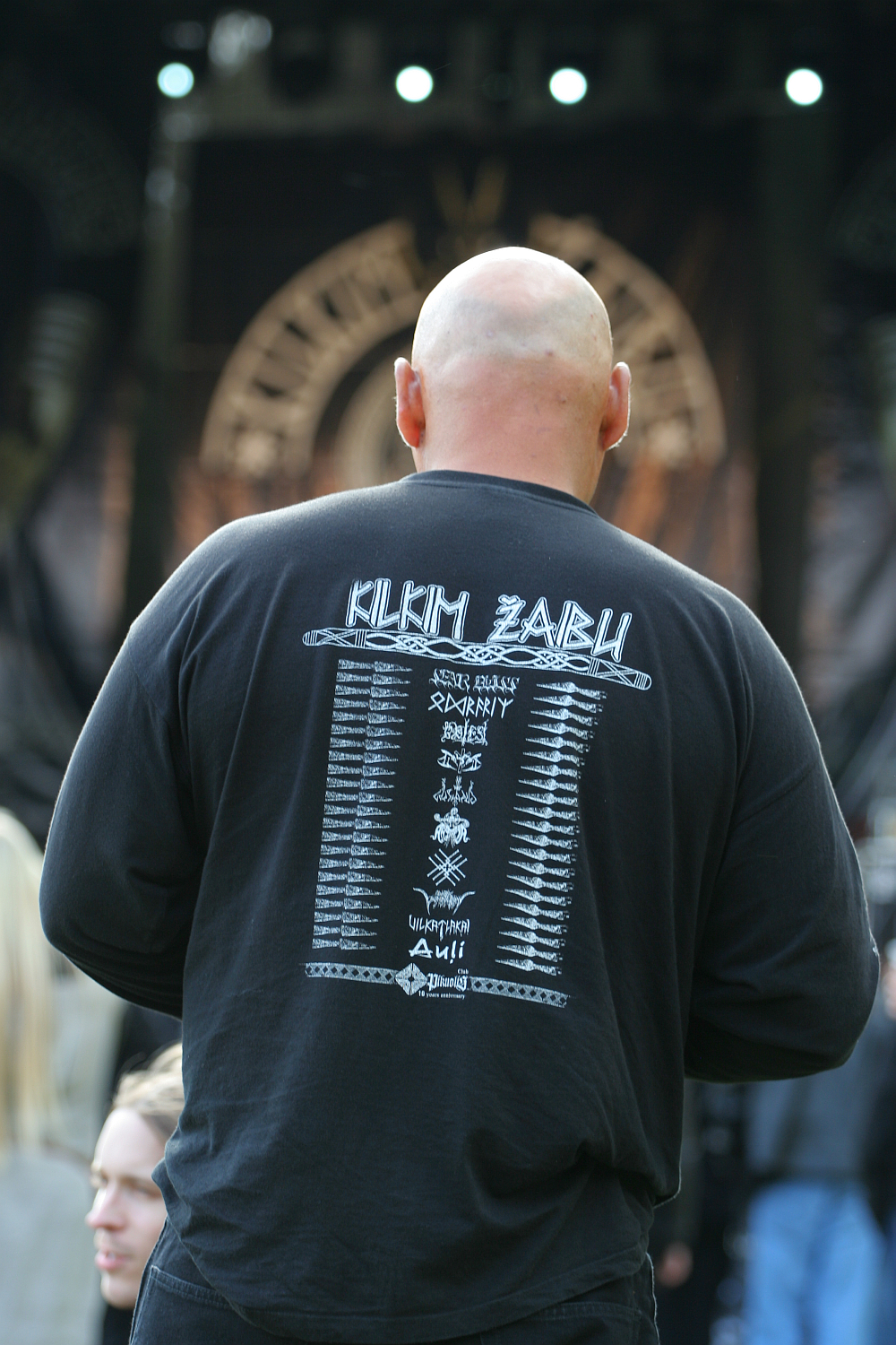 A bald man is seen from behind wearing a black long-sleeve shirt with white text featuring a list of band names and logos in a vertical arrangement. He is at an outdoor setting with a stage in the background, hinting at Kilkim Žaibu 2011. Other attendees are partially visible around him.