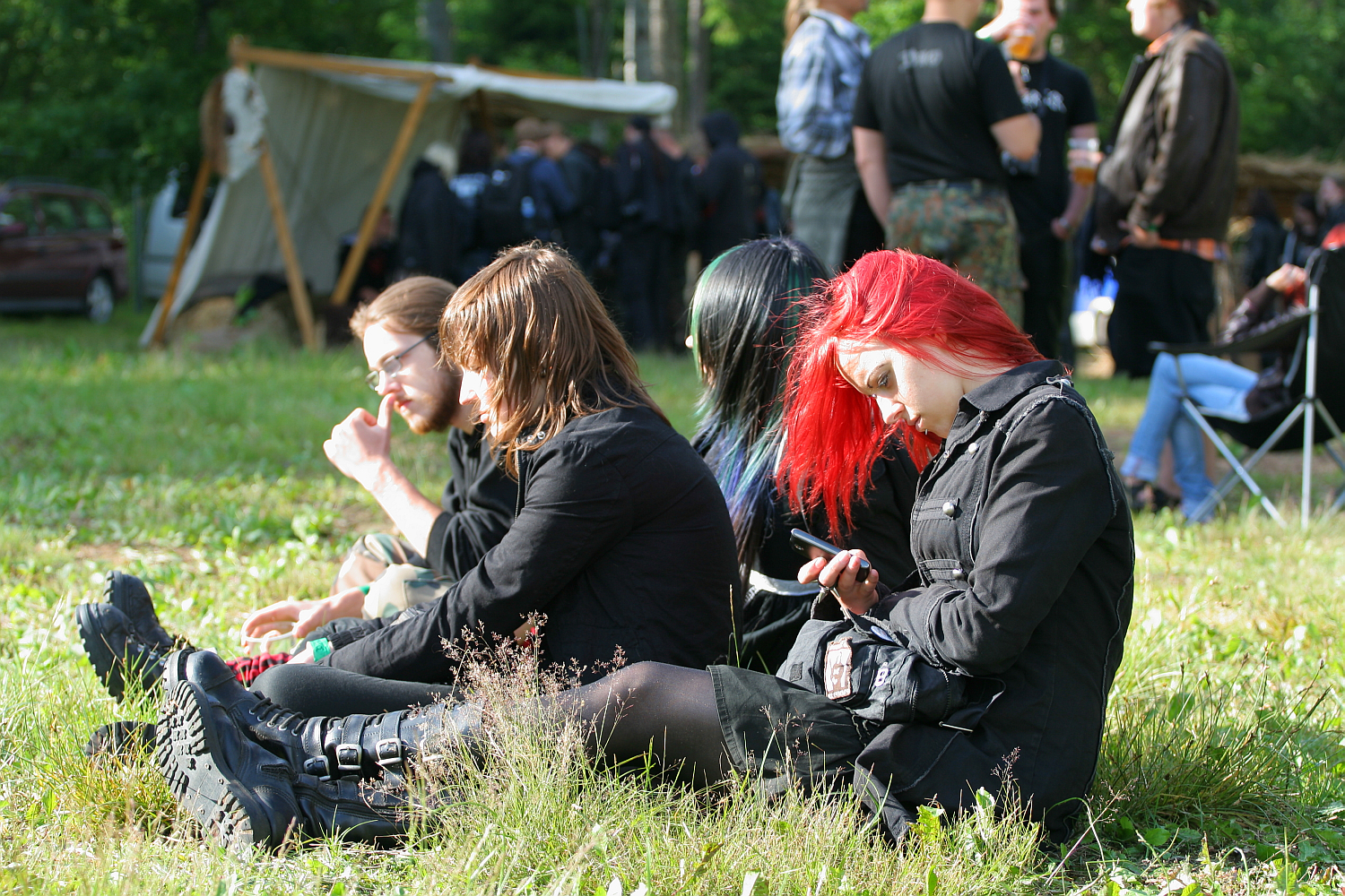 A group of four people dressed in black attire, including one with vibrant red hair, sit on low grass at the 2011 Kilkim Žaibu festival. One person examines a phone while others seem engaged in conversation and activities. In the background, more people are gathered near a fabric-covered shelter in an outdoor setting.