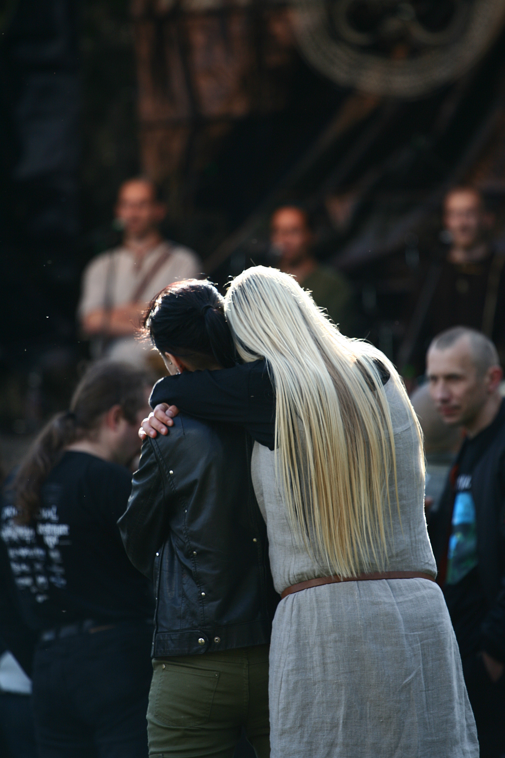 A person with long blonde hair wearing a grey outfit embraces another person with dark hair and a black jacket. They are standing in an outdoor festival setting, possibly Kilkim Žaibu 2011, with blurred individuals and equipment in the background. The atmosphere seems casual and social.