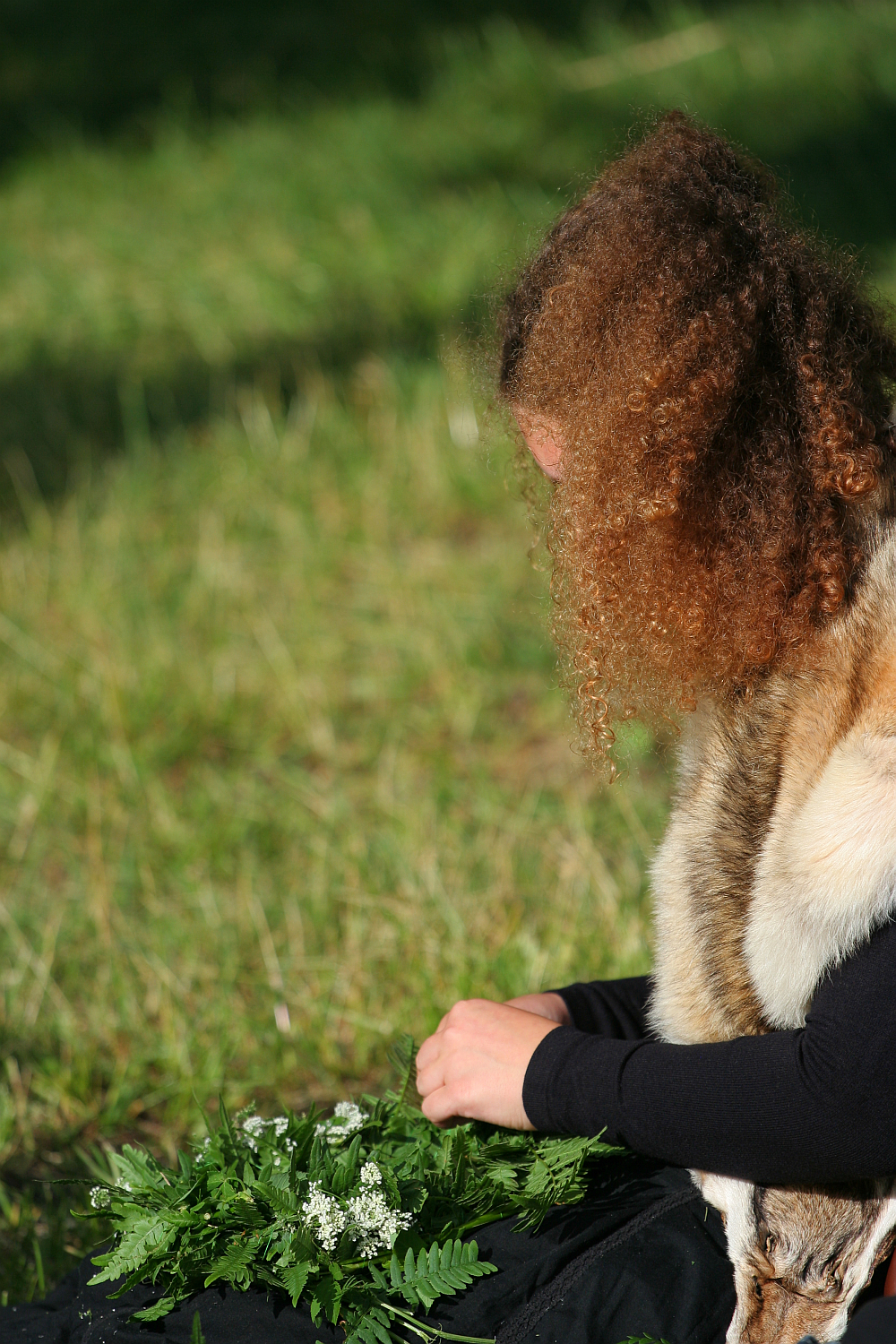 A person with curly brown hair, reminiscent of Kilkim Žaibu 2011, sits on the grass, focused on arranging green plants and white flowers in their hands. They are wearing a black long-sleeve shirt and a light brown fur shawl draped over their shoulders in the peaceful outdoor setting.