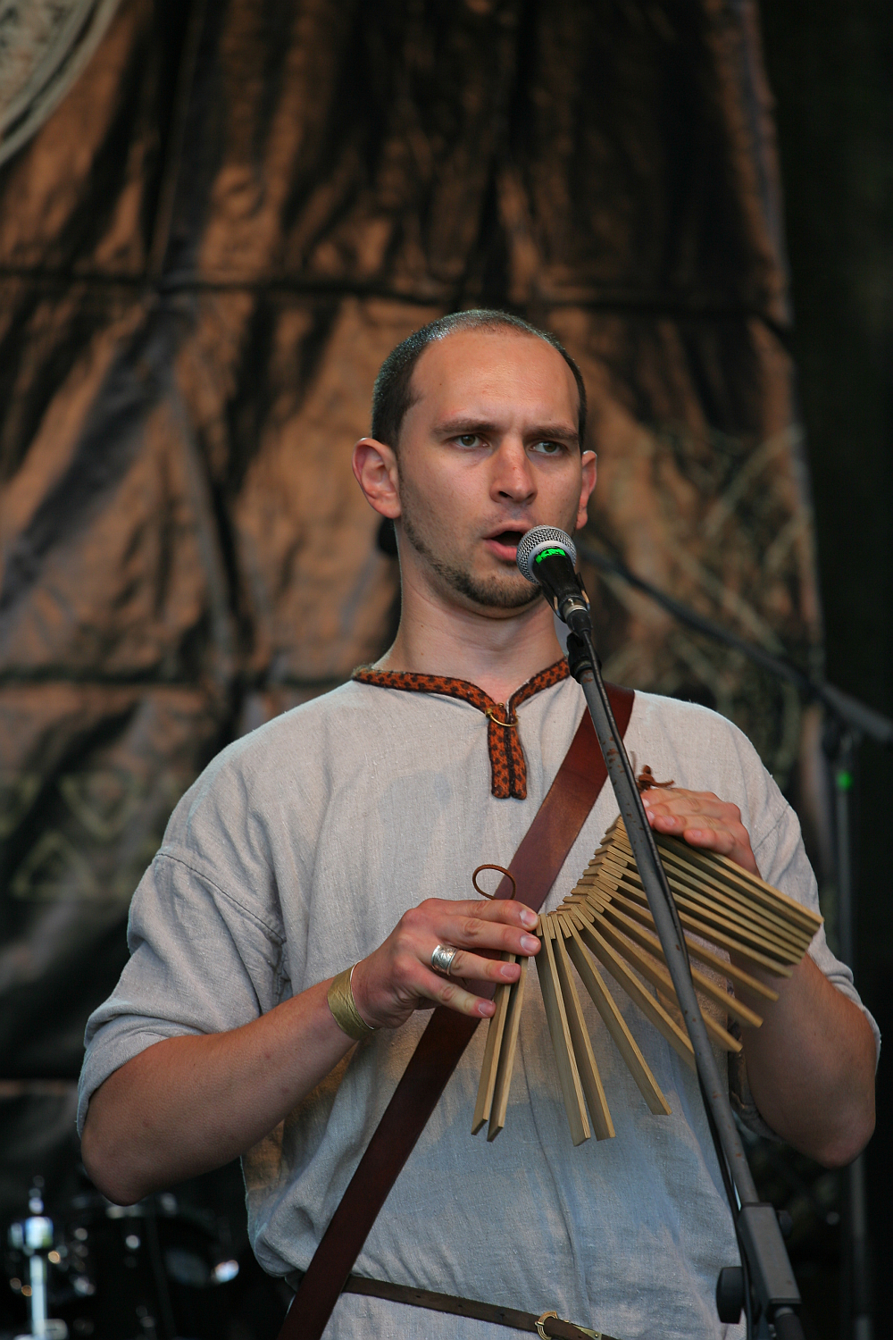 A person with short hair is playing a wooden reed instrument and singing into a microphone. They are wearing a light gray tunic with a brown tie neckline. The background features a dark draped cloth and stage lighting, indicating a live performance at the 2011 Kilkim Žaibu festival.
