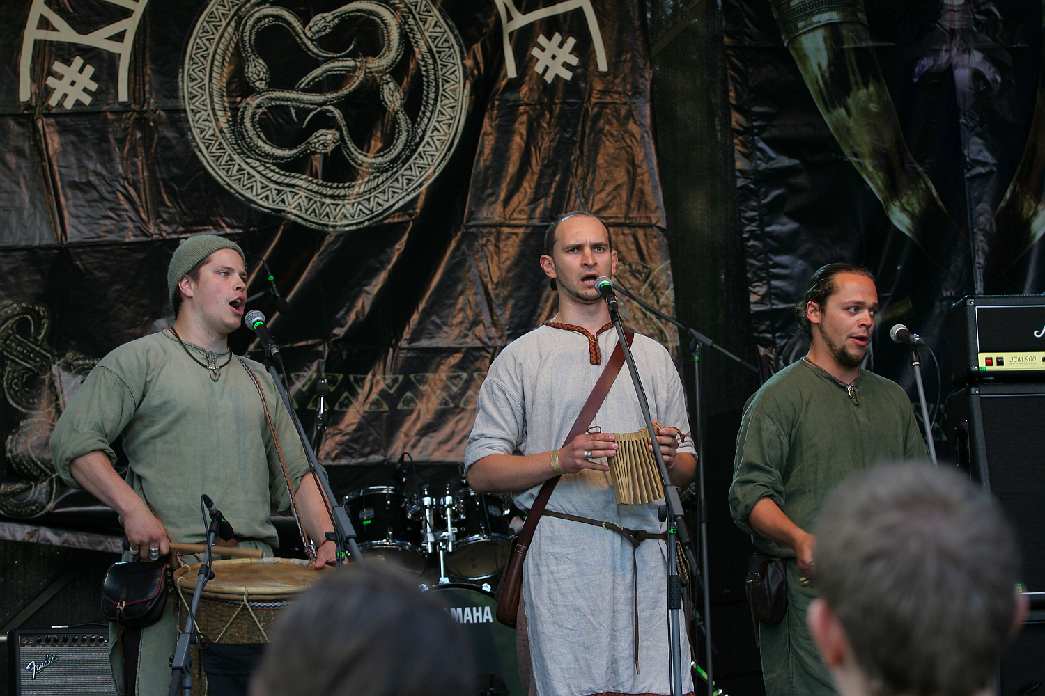 Three men dressed in medieval-style clothing perform on stage at Kilkim Žaibu 2011, each playing a different musical instrument. They are positioned in front of a large tapestry displaying a circular, intertwined snake design. One plays a drum, another holds a small box-shaped instrument, and the third has no visible instrument.