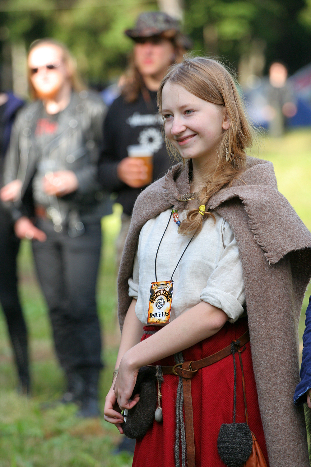 A young woman with blonde hair, wearing a beige shawl over a white shirt and red skirt, smiles outdoors at the 2011 Kilkim Žaibu festival. She has a pouch and a festival pass around her neck. Several people in the background, dressed casually, are out of focus. Trees and greenery are visible, indicating a nature setting.