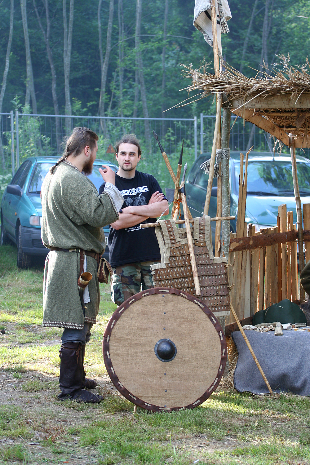 Two men are standing beside a medieval-style market stall with various wooden items at the Kilkim Žaibu festival in 2011. One man, in historical attire with a green tunic, brown belt, and boots, holds a cup. The other, in a black t-shirt and camouflage pants, stands with arms crossed. A round shield rests in front of the stall. Trees and parked cars