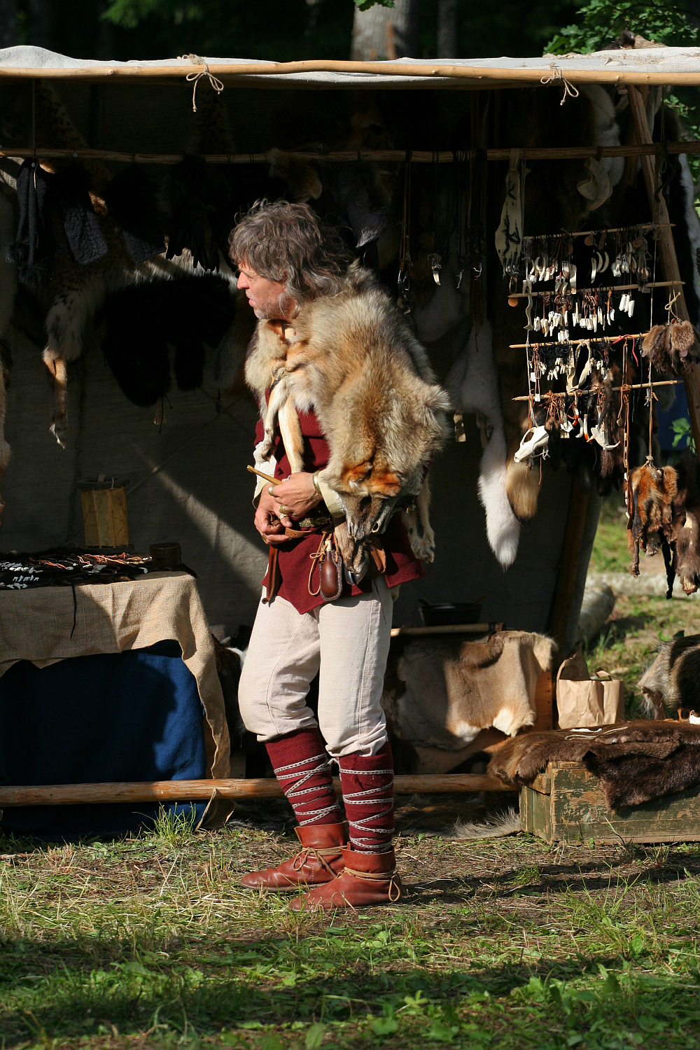 A man dressed in historical attire stands at an outdoor market. He wears a brown tunic, tan pants, and tall brown leather boots with red laces. Various animal pelts drape over his shoulders, and behind him, there are numerous animal pelts and handcrafted items displayed in a wooden stall at the 2011 Kilkim Žaibu festival.