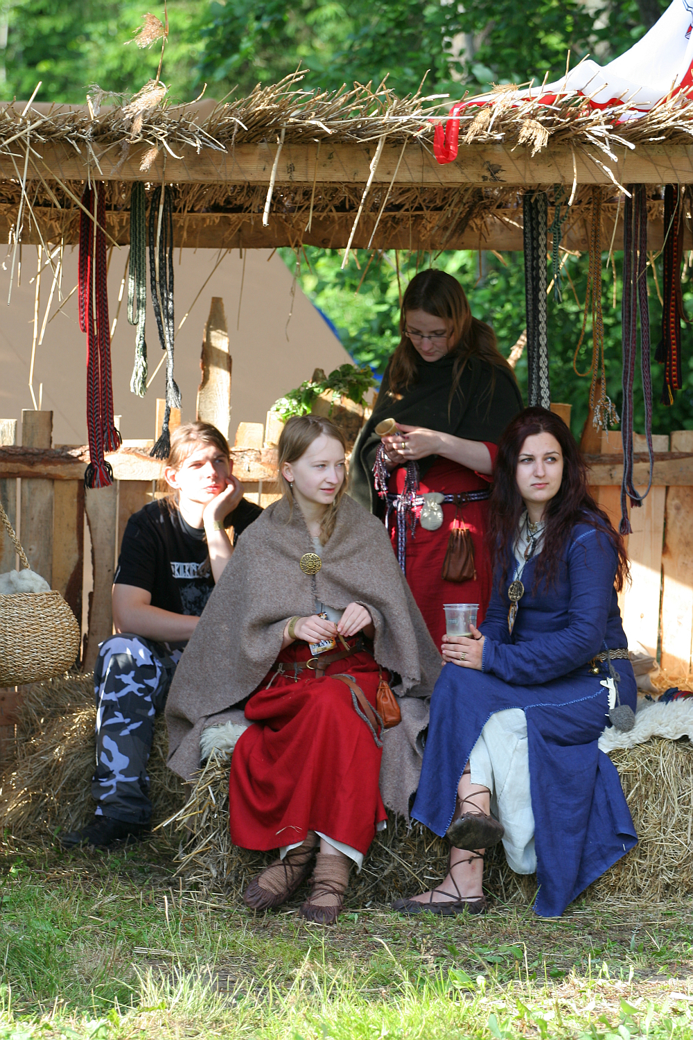 Four people in medieval costumes sit and stand in a rustic, open wooden shelter with a thatched roof at the 2011 Kilkim Žaibu festival. Two women in red and blue dresses sit on straw bales, one holding a drink. Another woman in red stands beside them while a man in camouflage pants sits nearby. Colorful ribbons hang in the background.