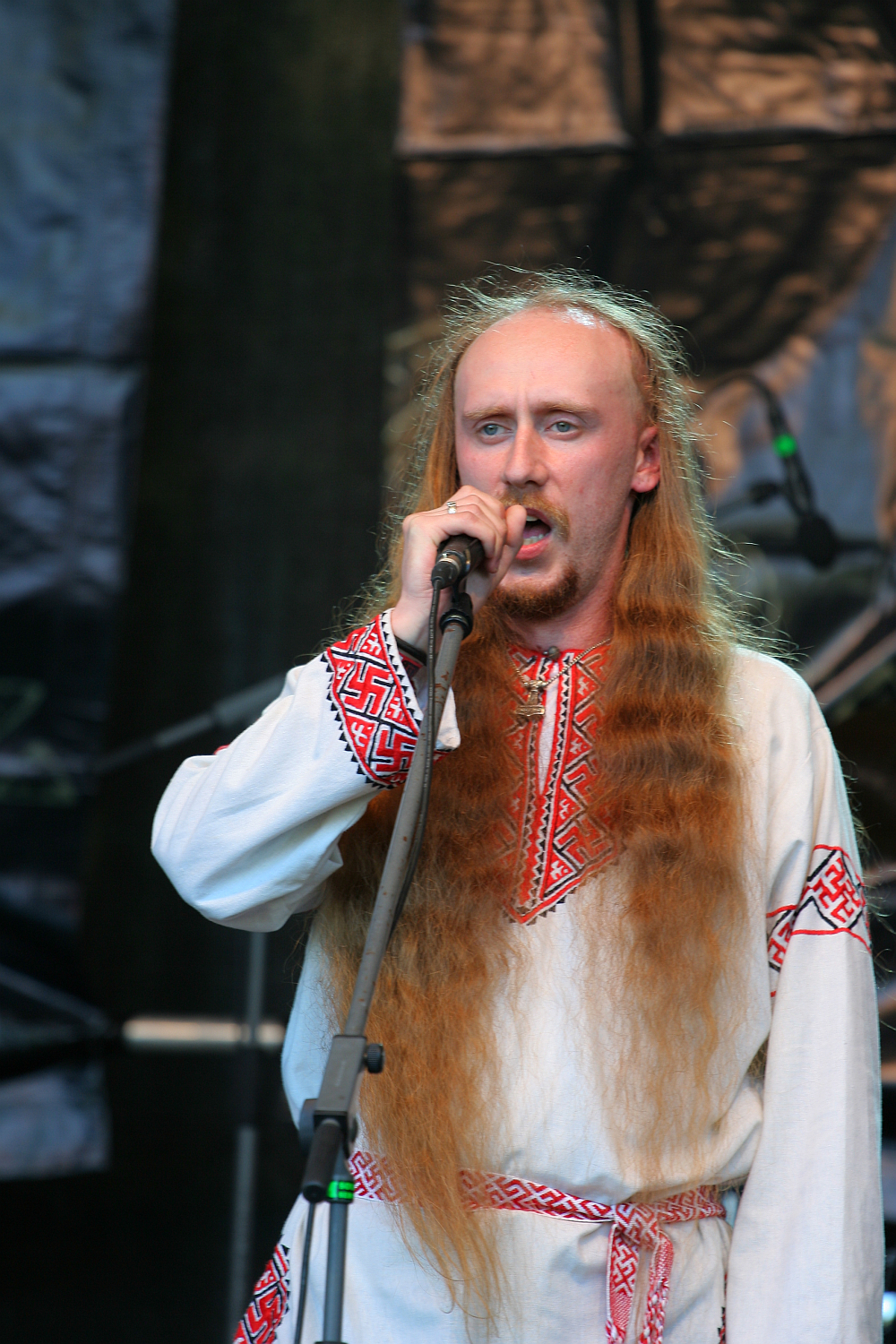 A man with long, red hair and a beard sings into a microphone. He is wearing a traditional, embroidered white shirt with red and black patterns around the collar and sleeves, reminiscent of styles seen at Kilkim Žaibu 2011. A matching embroidered belt completes his outfit. The background is dimly lit, focusing attention on the singer.