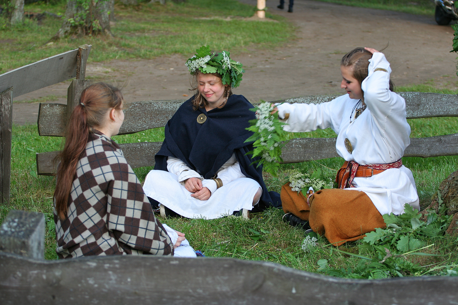 Three women in traditional clothing sit on the grass inside a wooden fence at the 2011 Kilkim Žaibu festival. One, wearing a flower crown, smiles with closed eyes. The second adjusts her hair, and the third has her back to the camera, wrapped in a plaid blanket. Leaves and branches lie scattered around them.