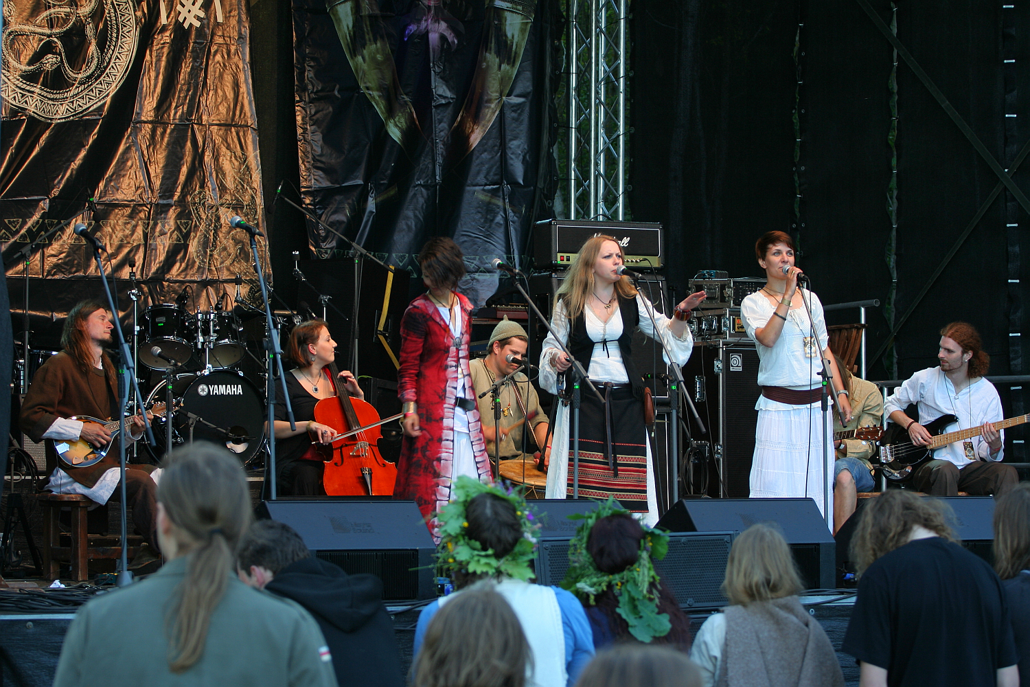 At the 2011 Kilkim Žaibu festival, a band performs on an outdoor stage, featuring members playing cello, guitars, and traditional instruments. Four vocalists, wearing various costumes including dresses and long coats, stand at the front of the stage. In the foreground, some audience members, one with a flower crown, watch the performance.