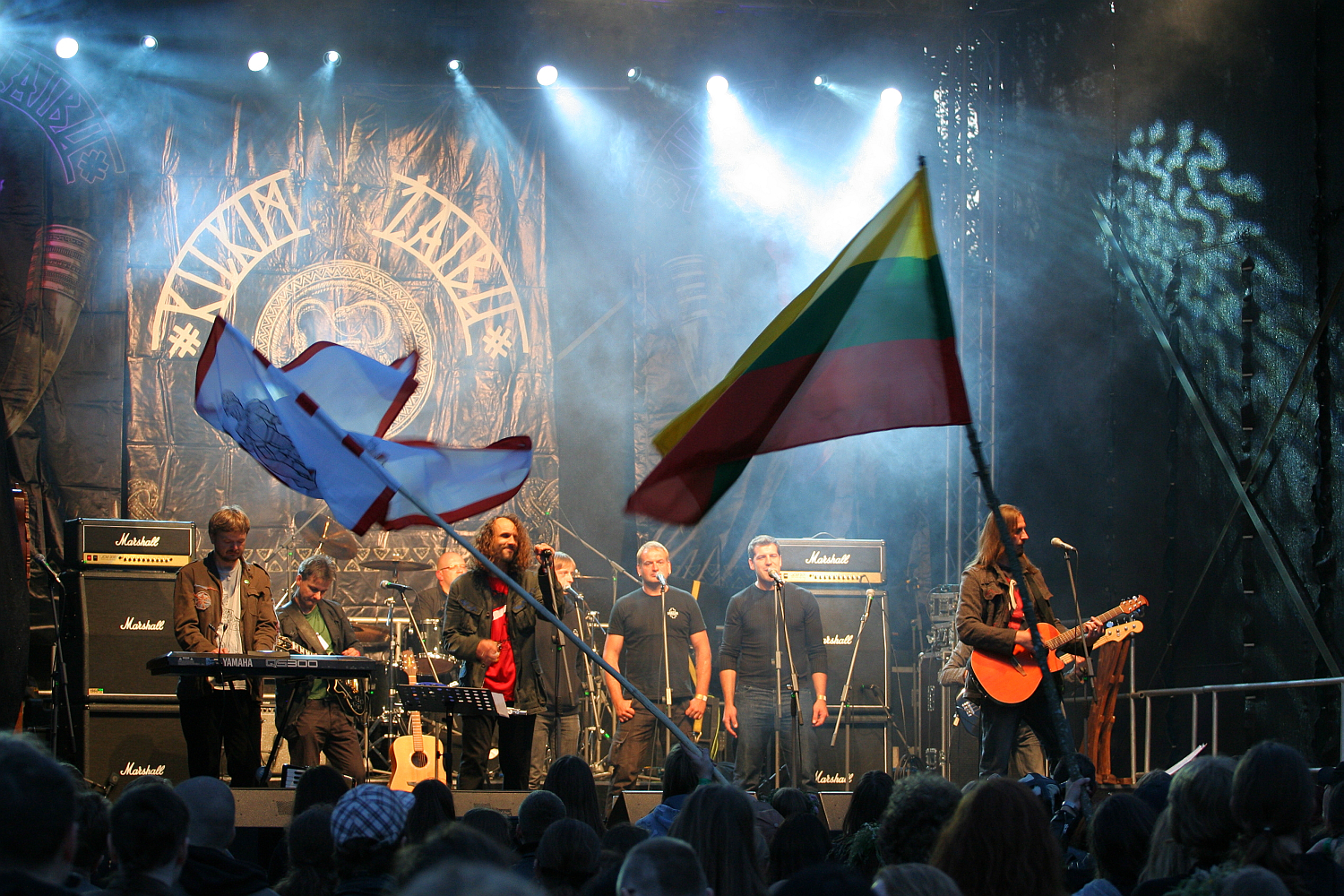A band performs on stage at an outdoor Kilkim Žaibu 2011 concert, with one musician playing an acoustic guitar. The stage backdrop features the band’s logo. Two large flags, one with Lithuanian colors and another with a different design, wave in the foreground. The audience watches the performance under stage lights.