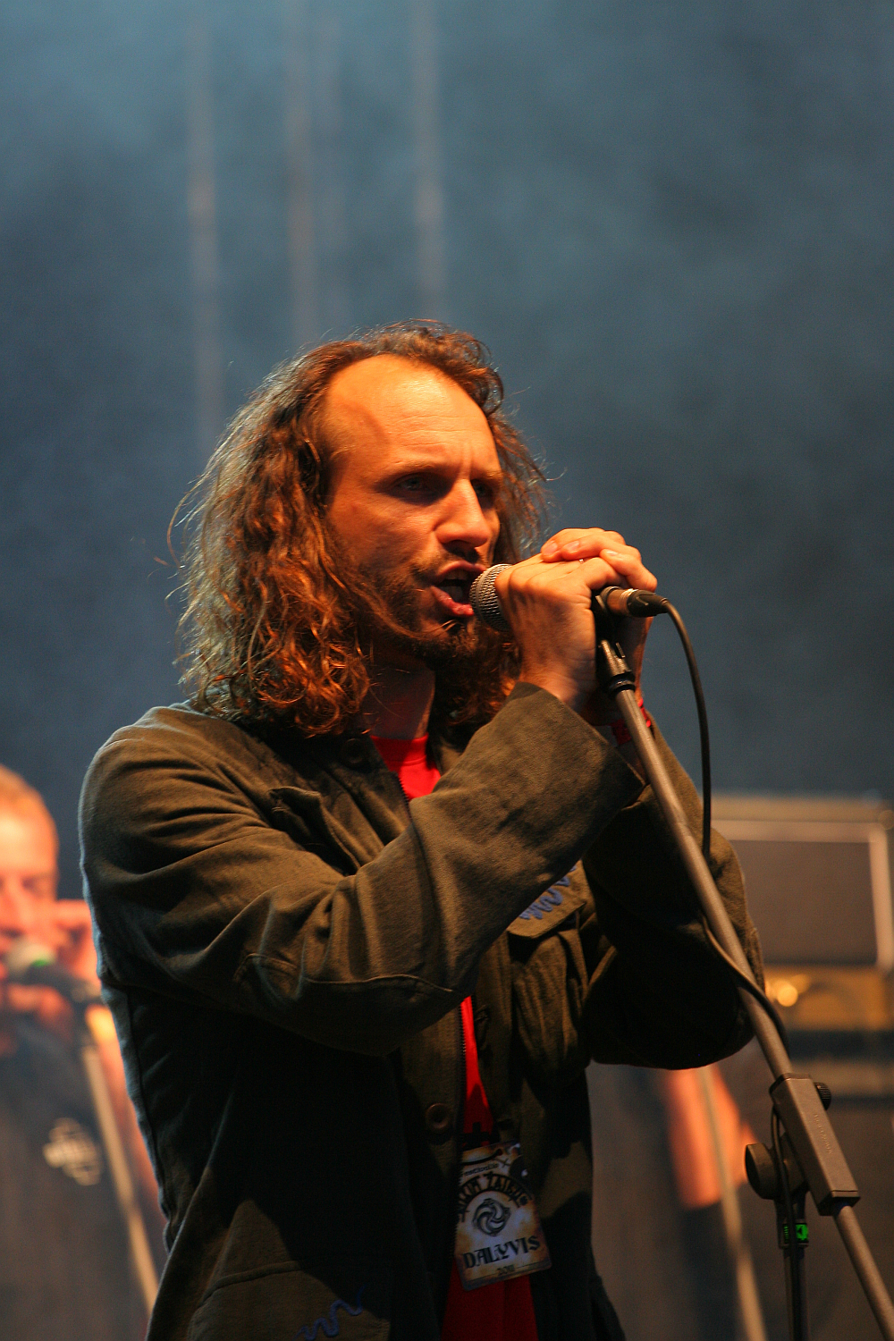 At the 2011 Kilkim Žaibu festival, a man with long curly hair performs on stage, passionately singing into a microphone he holds with both hands. He wears a dark jacket over a red shirt and has a lanyard around his neck. The background is dark with heavy stage lighting and another person, partially visible, is in the background.