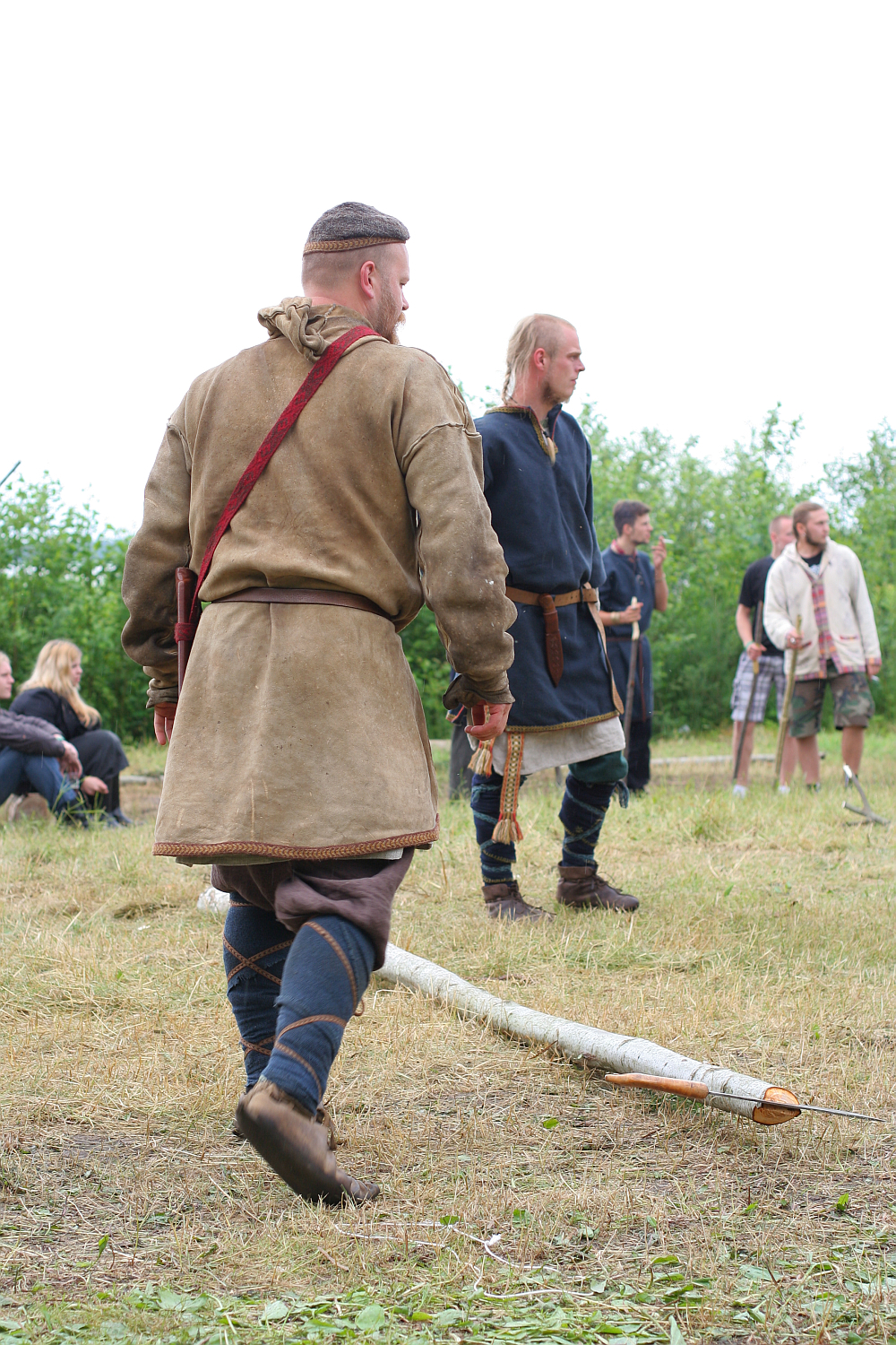 Two men dressed in historical medieval attire participate in an outdoor event, reminiscent of Viking-era clothing. Set in a grassy, open area during Kilkim Žaibu 2011, one man walks past a fallen wooden branch while others linger in the background. Spectators are seated comfortably in folding chairs.