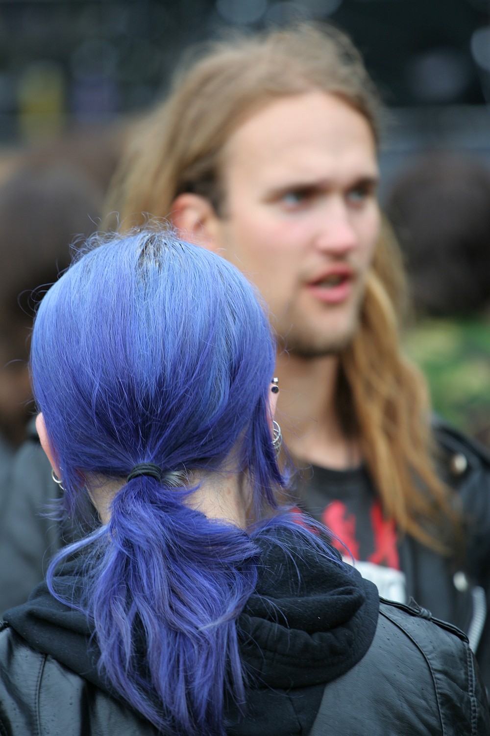 A person with long, vibrant blue hair tied in a ponytail is in the foreground, facing away. In the background, slightly out of focus, stands another with long blonde hair and a beard. Both are outdoors at Kilkim Žaibu 2011, wearing dark clothing. The scene suggests a casual or social setting.