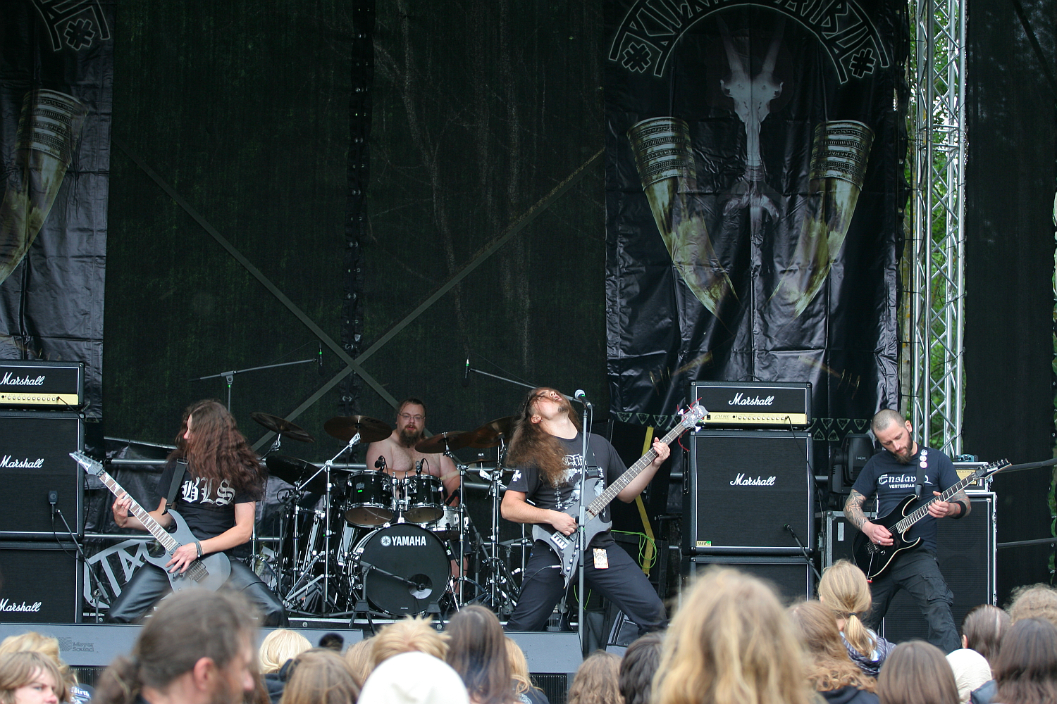 A metal band is performing on the outdoor stage of Kilkim Žaibu 2011, adorned with large, dark banners. Four members, dressed in black, are playing electric guitars and bass, with long hair flying as they headbang. The drummer is visible at the back, surrounded by Yamaha drums. The audience watches intently with some raised hands.