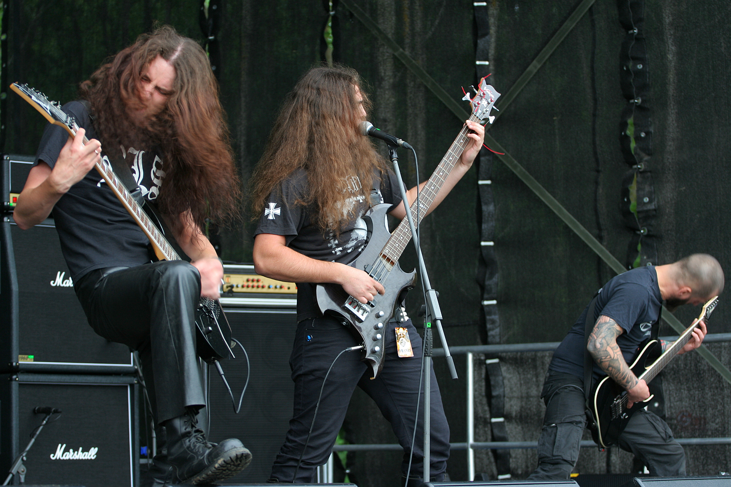 Three long-haired metal musicians perform energetically on stage at the 2011 Kilkim Žaibu festival. The lead singer, with both hands on his guitar, sings into a microphone, while the flanking guitarists headbang intensely. Amplifiers, including Marshall stacks, are visible in the background. The overall scene is dynamic and intense.