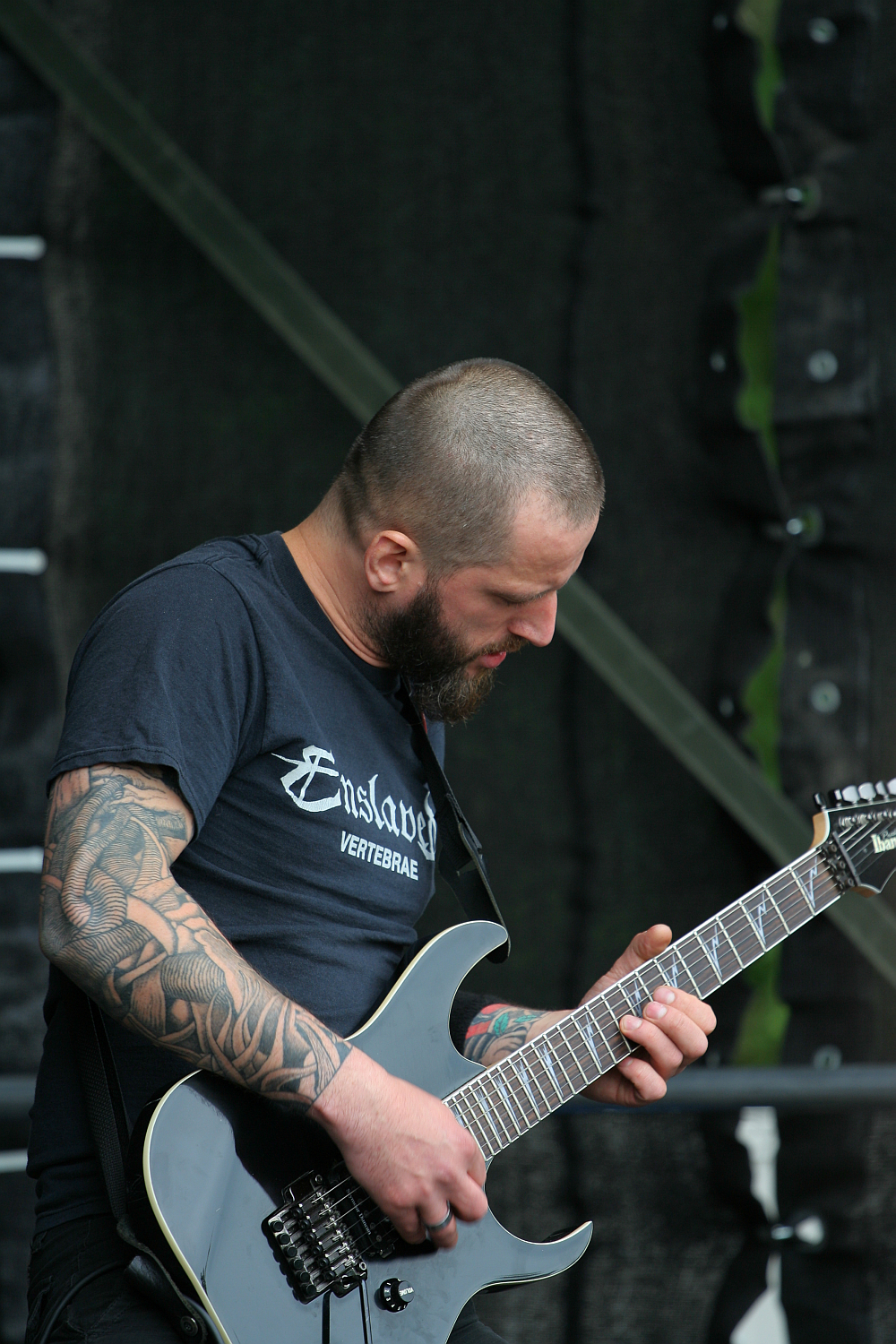 A bearded man with a shaved head and intricate tattoos on his left arm plays an electric guitar. Wearing a black t-shirt with white text, he is focused on his instrument. The background shows a dark, partially blurred stage setup, reminiscent of Kilkim Žaibu 2011.