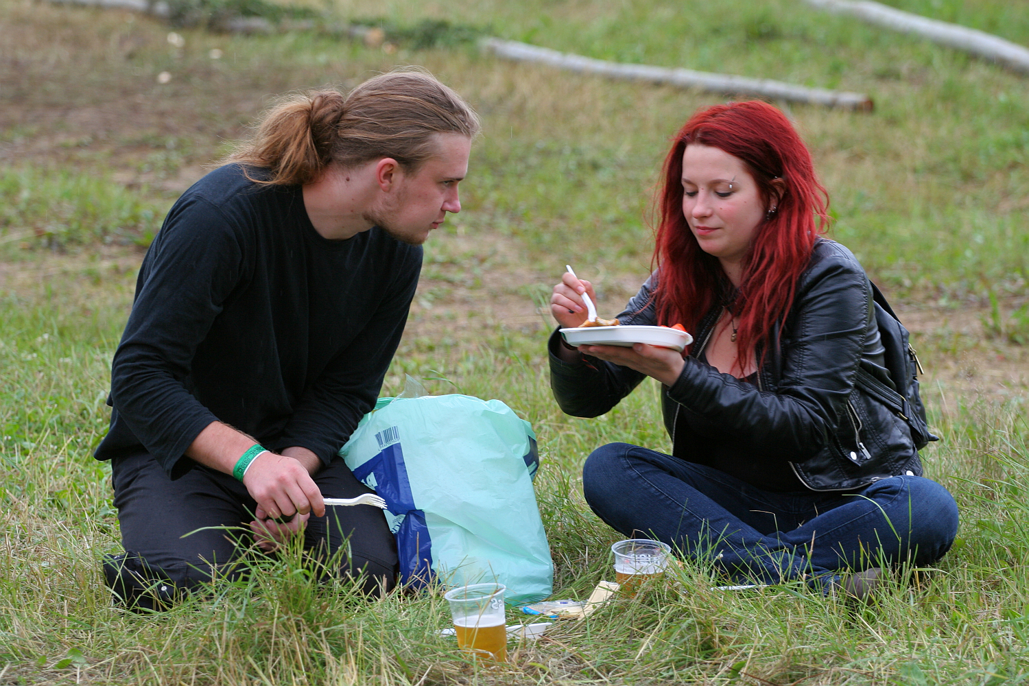 Two people, a man with long hair tied back and a woman with red hair, sit on grass sharing a meal at Kilkim Žaibu 2011. The woman, wearing a black leather jacket, holds a plate and fork while the man looks on. They have drinks in plastic cups on the ground next to them, along with a plastic bag and utensils.