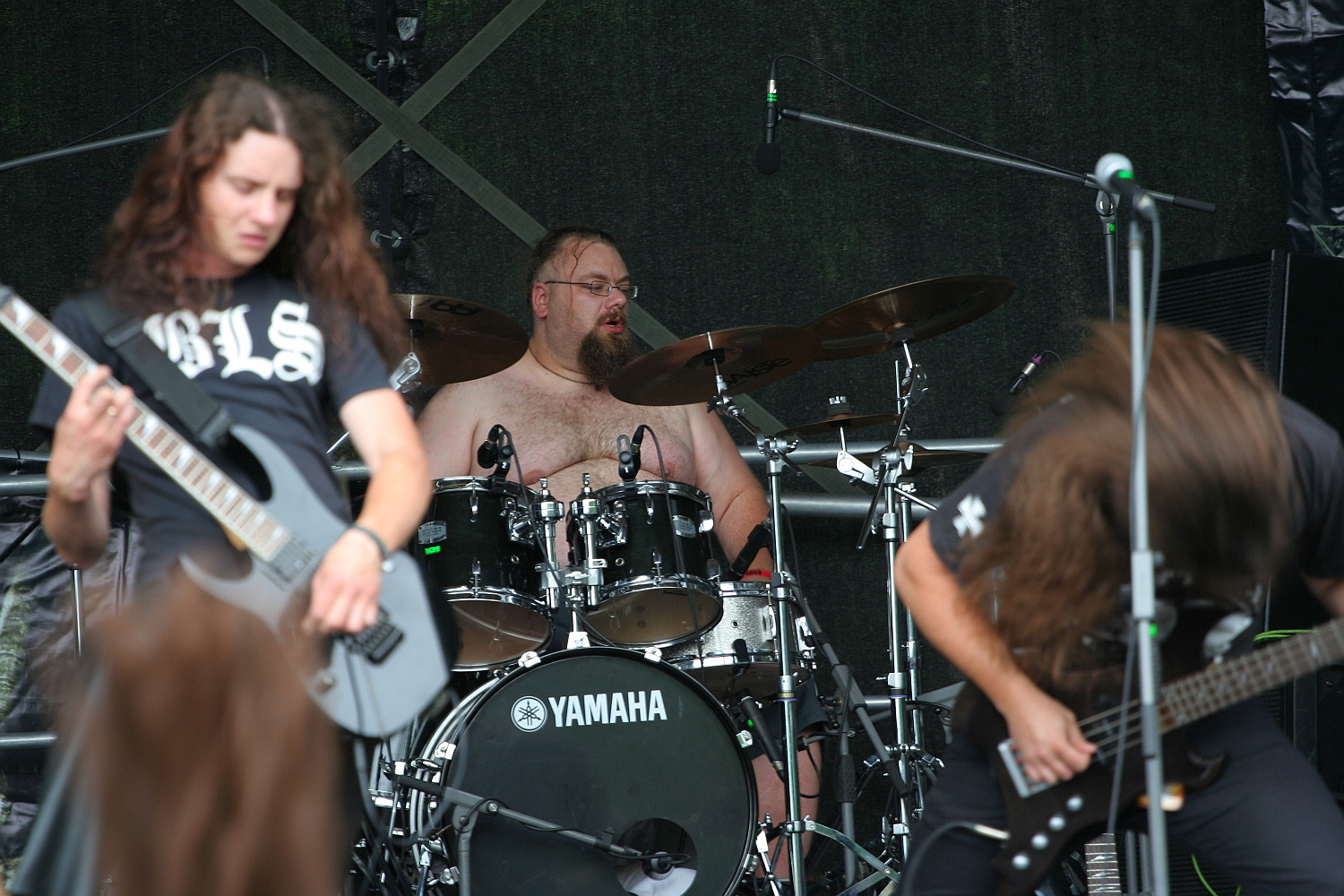 A rock band performs on stage at Kilkim Žaibu 2011. The drummer, shirtless, passionately plays on a Yamaha drum kit. Two guitarists with long hair are in the foreground, intensely engaged with their instruments, headbanging. The stage backdrop is dark, emphasizing the musicians' energetic presence.
