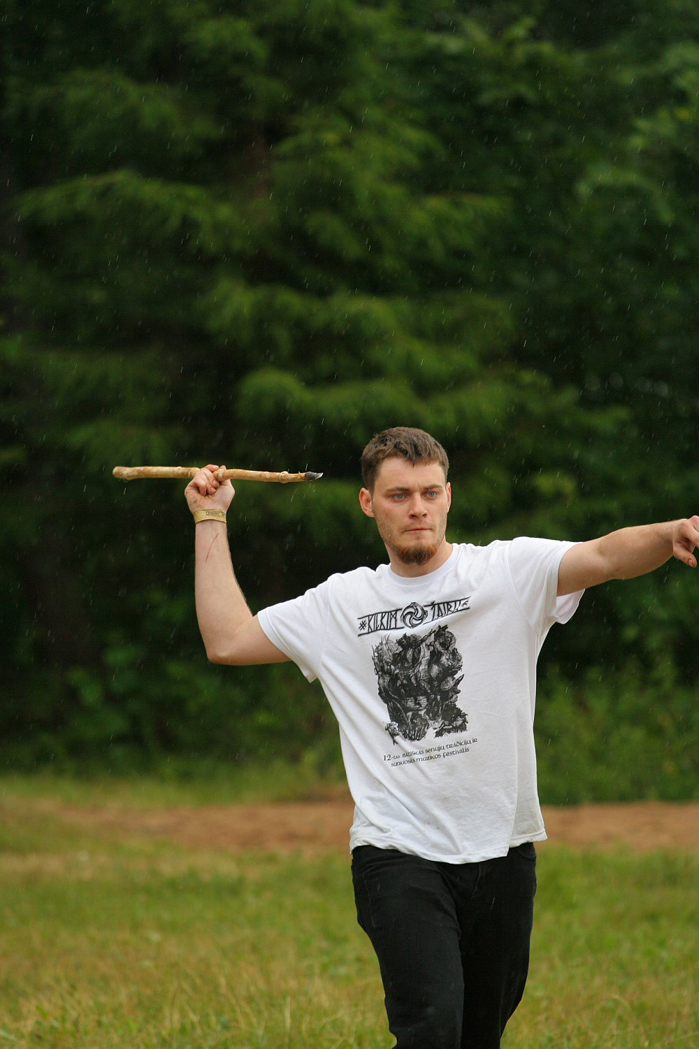 A person with short hair wearing a white graphic T-shirt and dark pants stands on grassy terrain, raising one arm to throw a stick and extending the other arm forward. There are trees in the blurred background, and the weather appears to be slightly misty or rainy, reminiscent of a scene from Kilkim Žaibu 2011 festival.