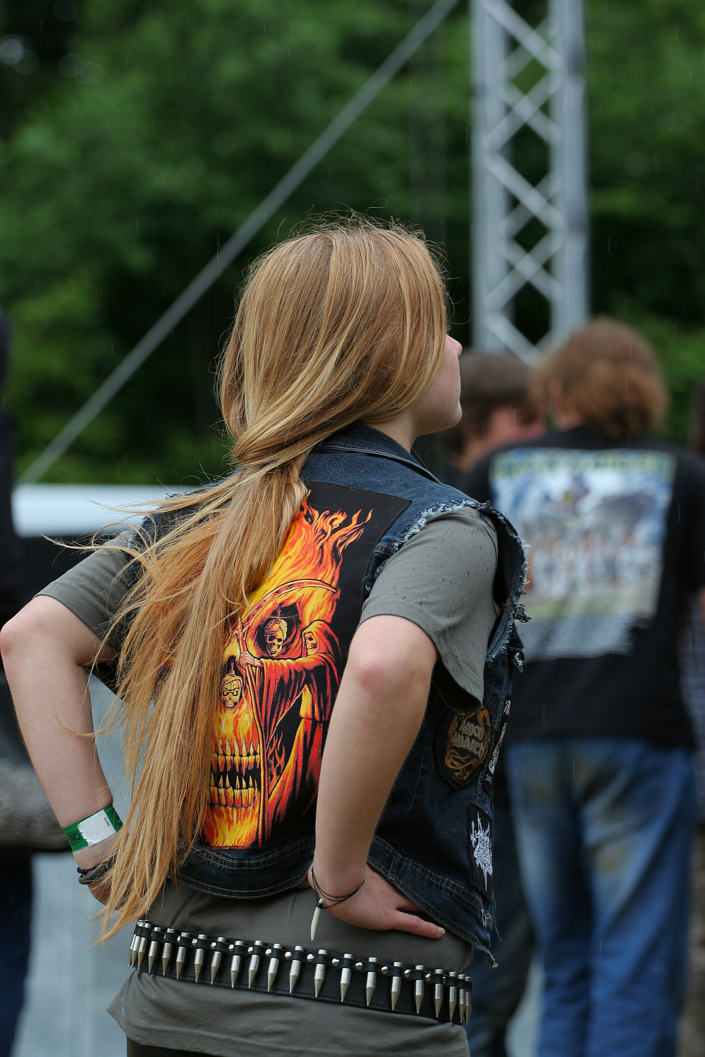 A person with long blonde hair is facing away from the camera at an outdoor event, possibly Kilkim Žaibu 2011. They are wearing a black denim vest featuring a graphic of a flaming skull and a grey t-shirt. A studded belt can be seen at their waist. Other event attendees are visible in the background.
