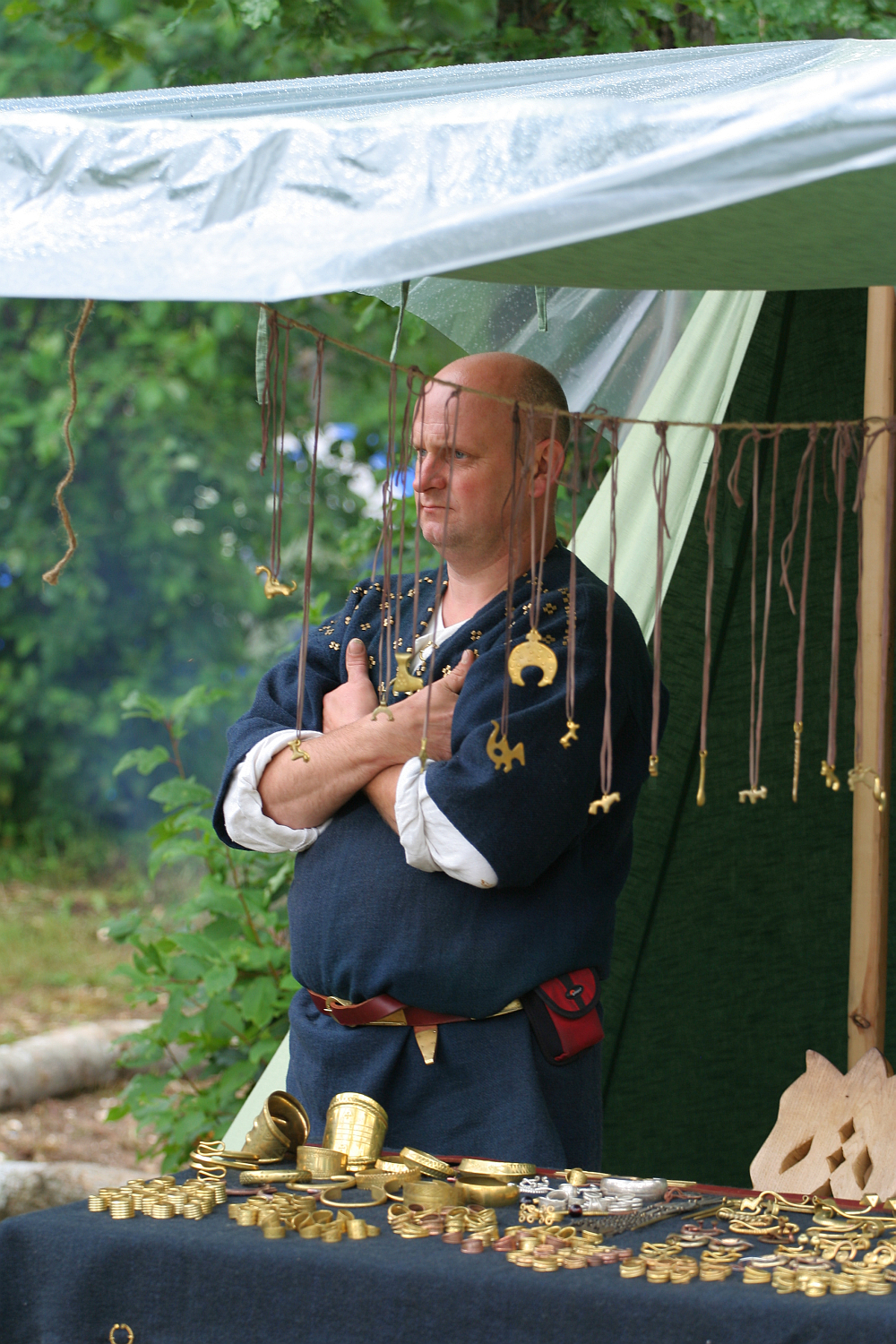 A person in medieval attire stands under a canopy with arms crossed. Various brass artifacts, including keys and coins, hang on strings in front. The table displays more artifacts on a black cloth. The setting appears to be the 2011 Kilkim Žaibu festival, with green foliage in the background.